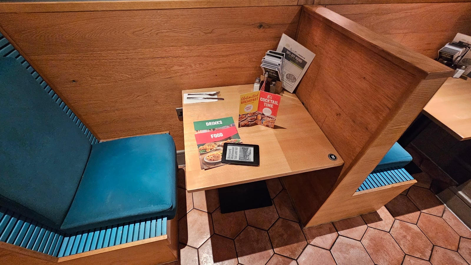 A booth with teal seats, a wooden table set with utensils, menus, a Kindle device, and condiments against a wood-paneled wall on a hexagon-tiled floor.