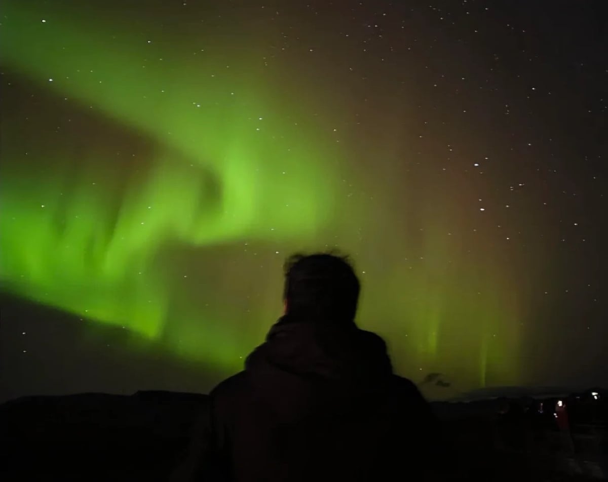 A person stands silhouetted, watching vivid green northern lights dance across a starry night sky.
