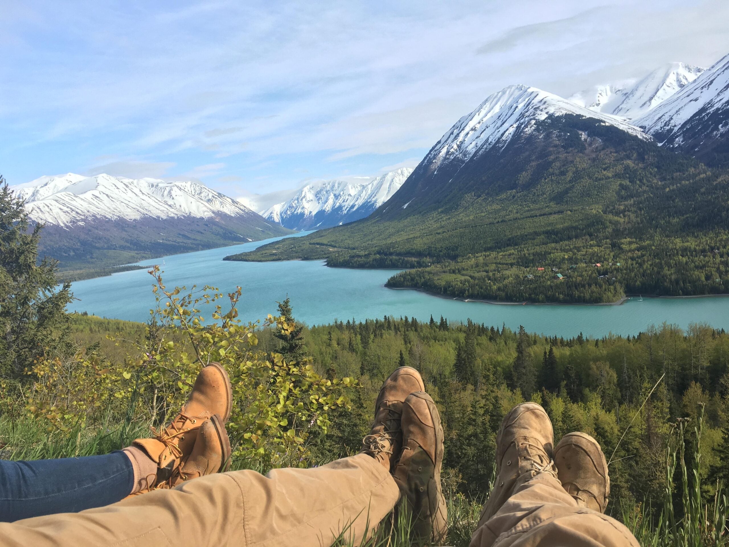Two hikers wearing boots relax on grass, overlooking a turquoise river winding through a forested valley with snow-capped mountains under a partly cloudy sky—a perfect moment captured in photos and posted to share their adventure.