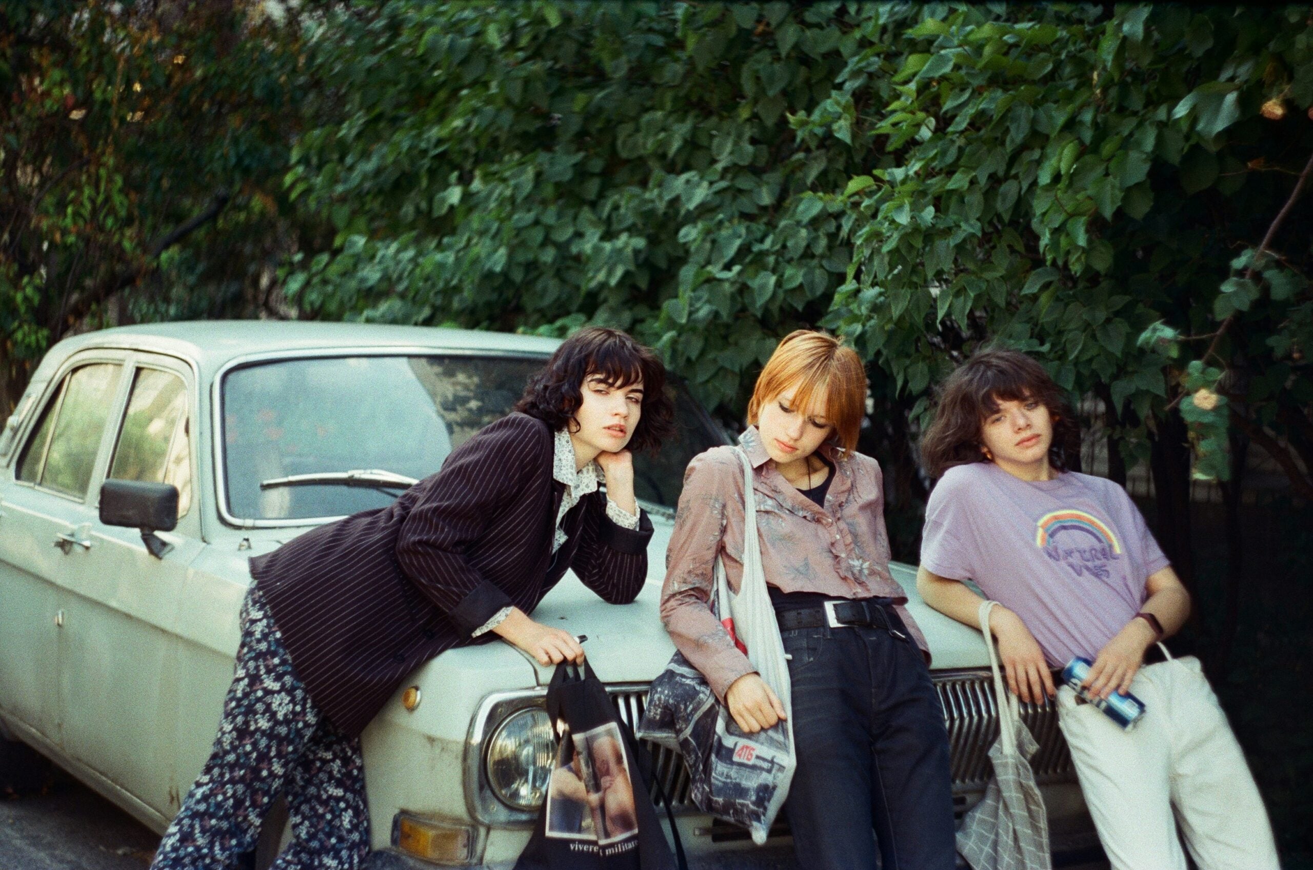 Three young people lean casually against an old, light-colored car parked by green trees. They wear vintage-style clothing and relaxed expressions, each holding a bag or item in their hands.