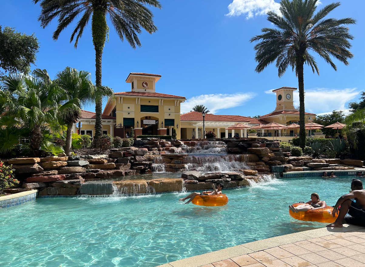 People relax on orange float tubes in a resort pool with a waterfall, surrounded by palm trees and a large, yellow resorts building under a blue sky.