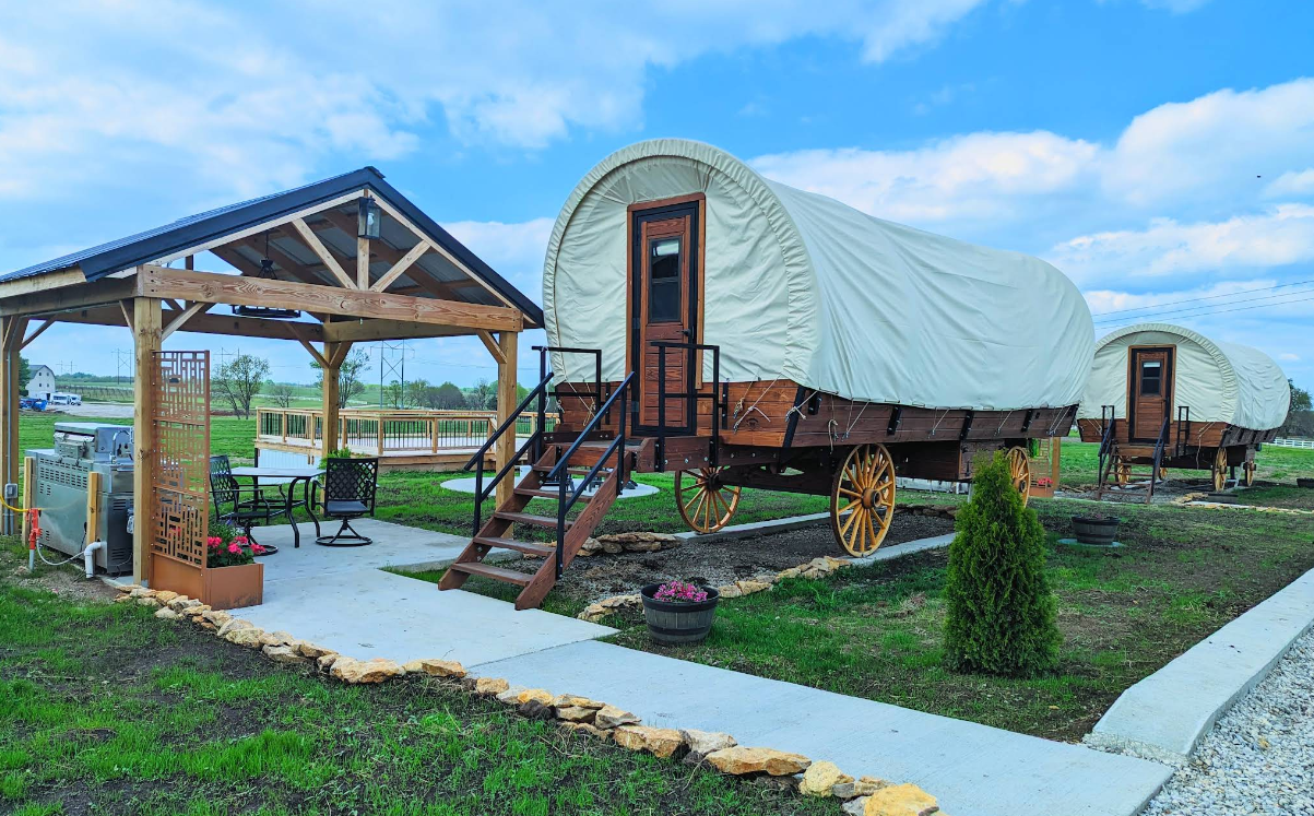 Two covered wagons converted into charming cabins sit on a grassy plot, each with stairs and wooden wheels. Reminiscent of cozy resorts, a small pavilion with chairs and a table is nearby under a partly cloudy blue sky.
