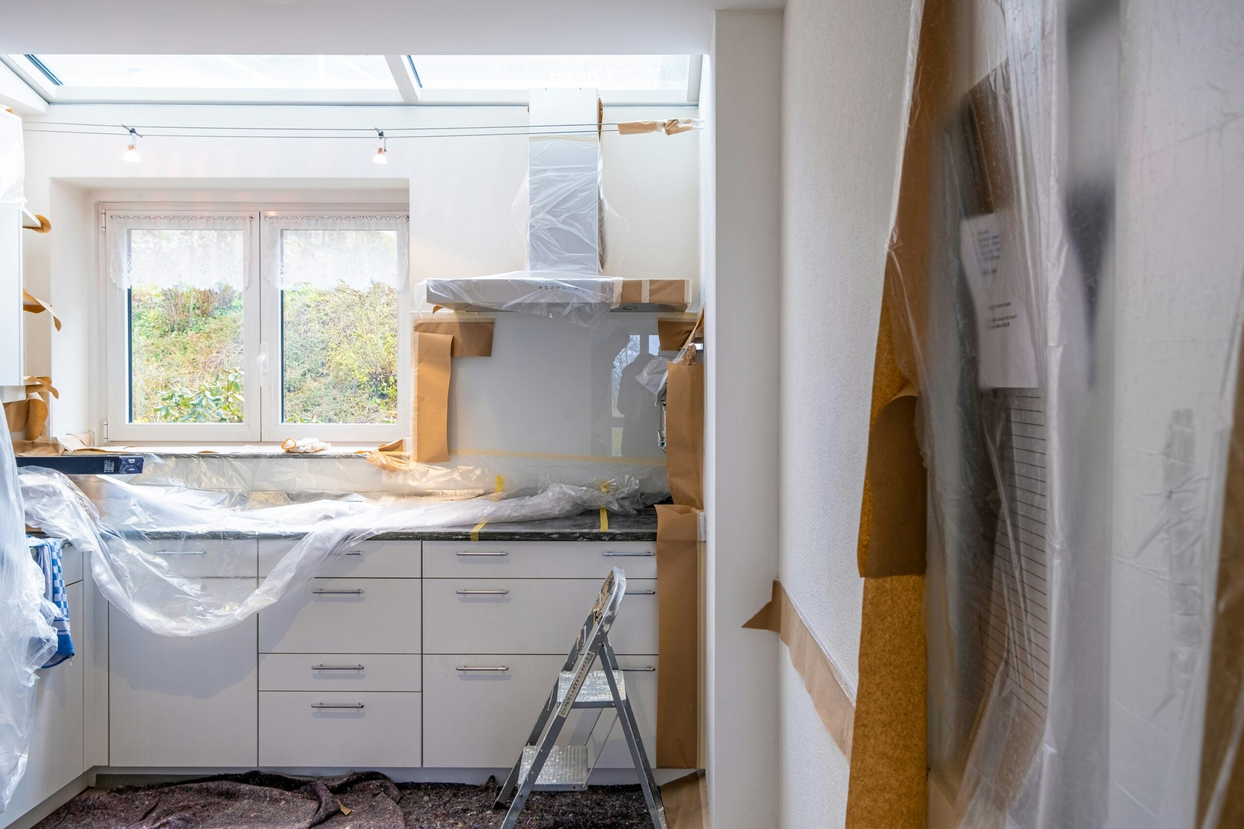 A kitchen under renovation with cabinets, countertop, and appliances covered in plastic sheets. A ladder stands near the counter, and natural light comes in from a window above the sink.