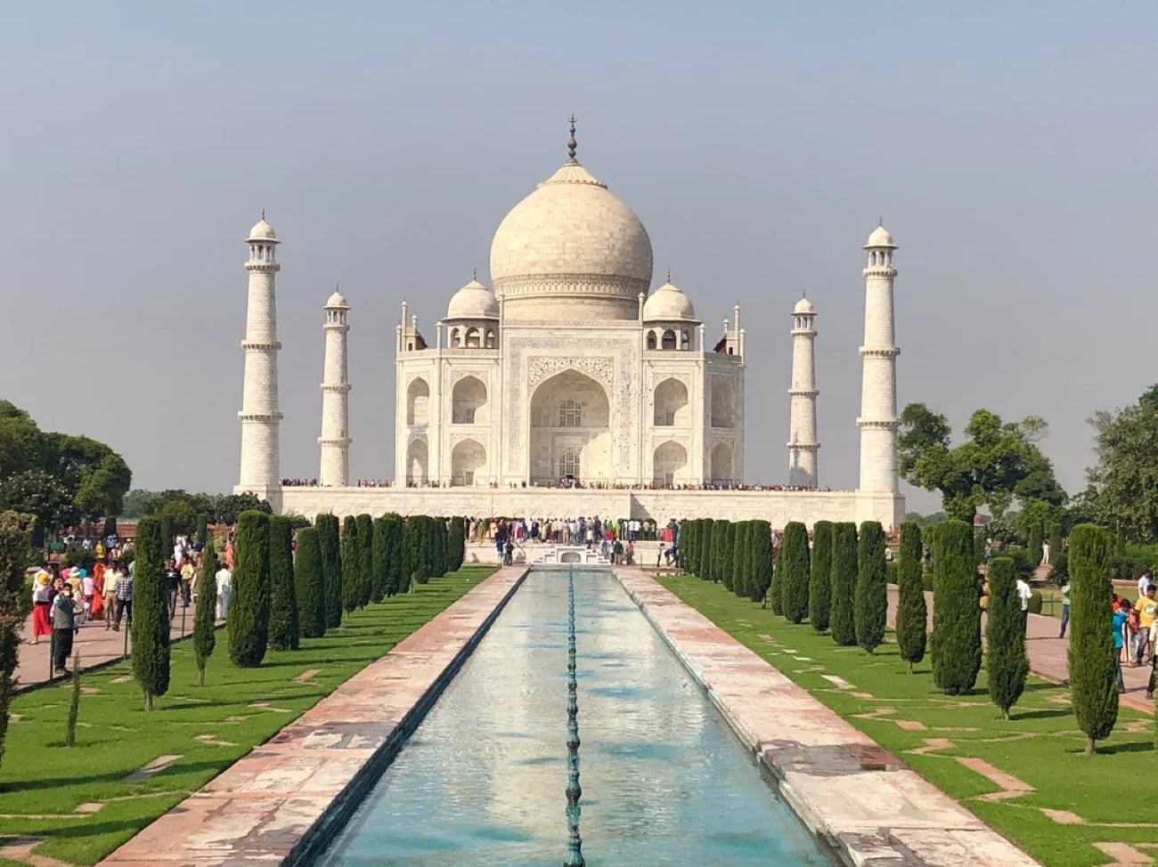 The Taj Mahal in India, with its white marble dome and minarets, stands in the background—a breathtaking sight nestled within one of the Biggest Cities In The World. A long reflecting pool leads to the entrance, flanked by green gardens, under a clear blue sky. Tourists are visible around the monument.