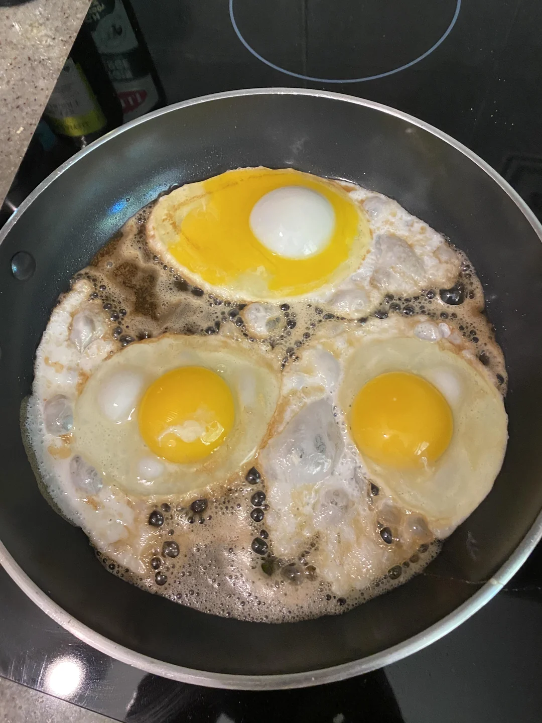 Three eggs are frying in a black pan; two have visible yolks, while the third has a white, opaque yolk. The eggs are surrounded by bubbling oil or butter on a stovetop.