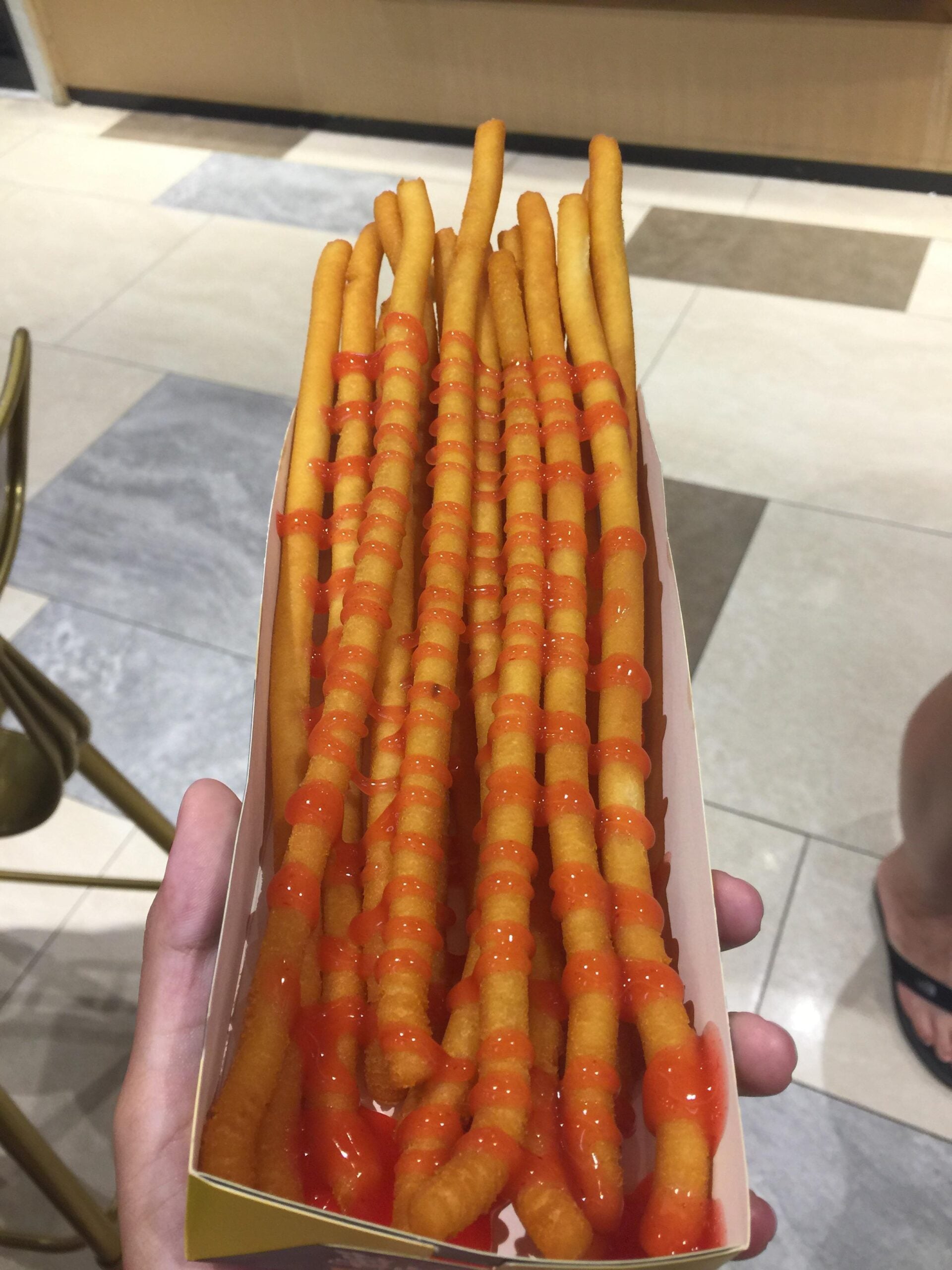 A hand holds a tray of long, thin fried snacks topped with a zigzag of orange sauce. The background shows tiled flooring and part of a chair.