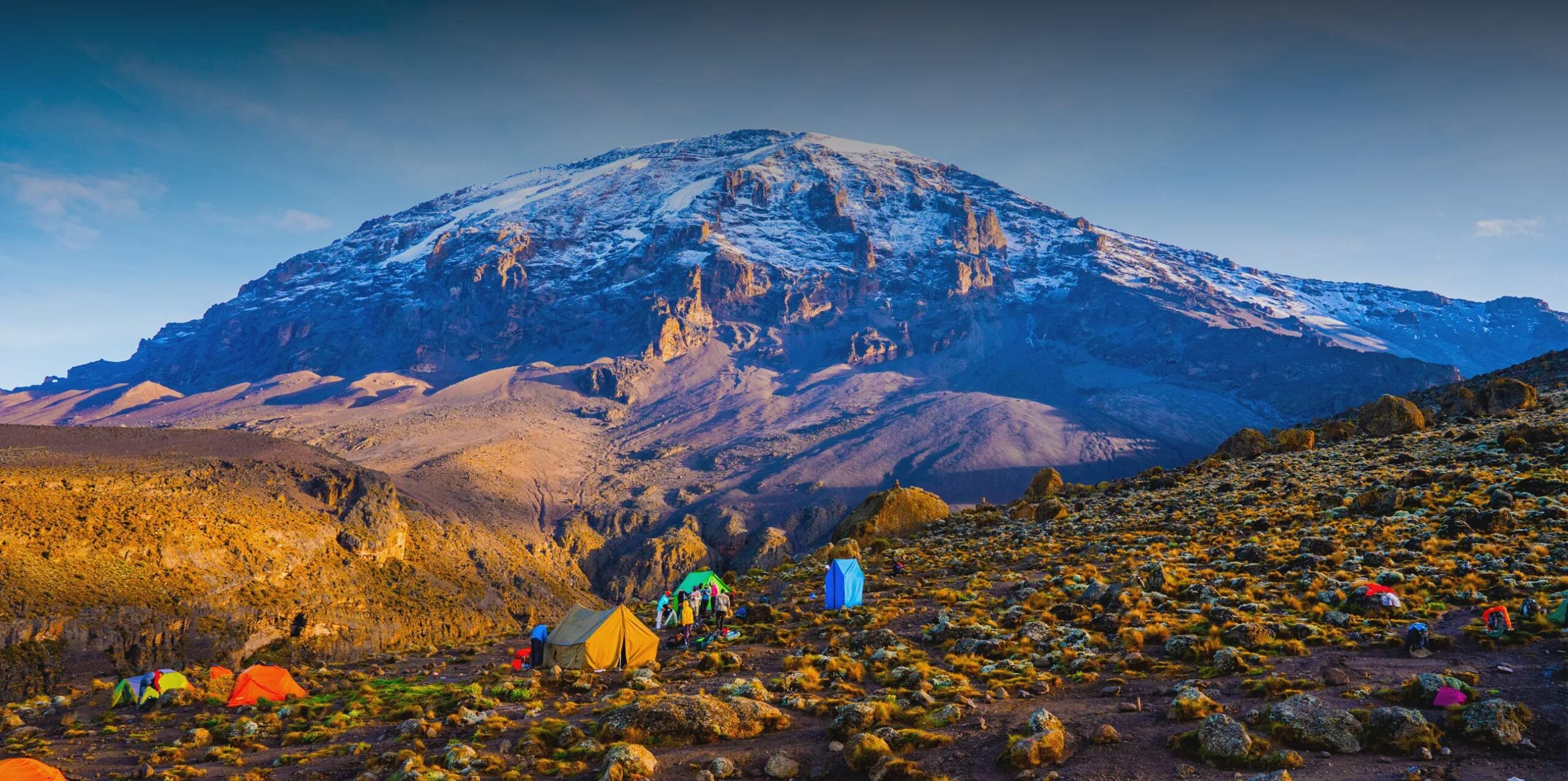Colorful tents are pitched on rocky ground in front of Mount Kilimanjaro, topped with snow and illuminated by sunlight, under a clear blue sky.