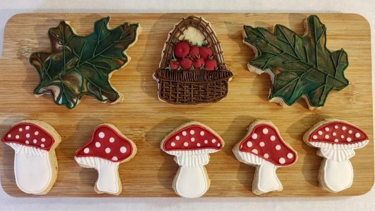 Cookies shaped like mushrooms, oak leaves, and a basket of red apples are arranged on a wooden board. These seasonal cookies are decorated with colorful icing to resemble real objects.