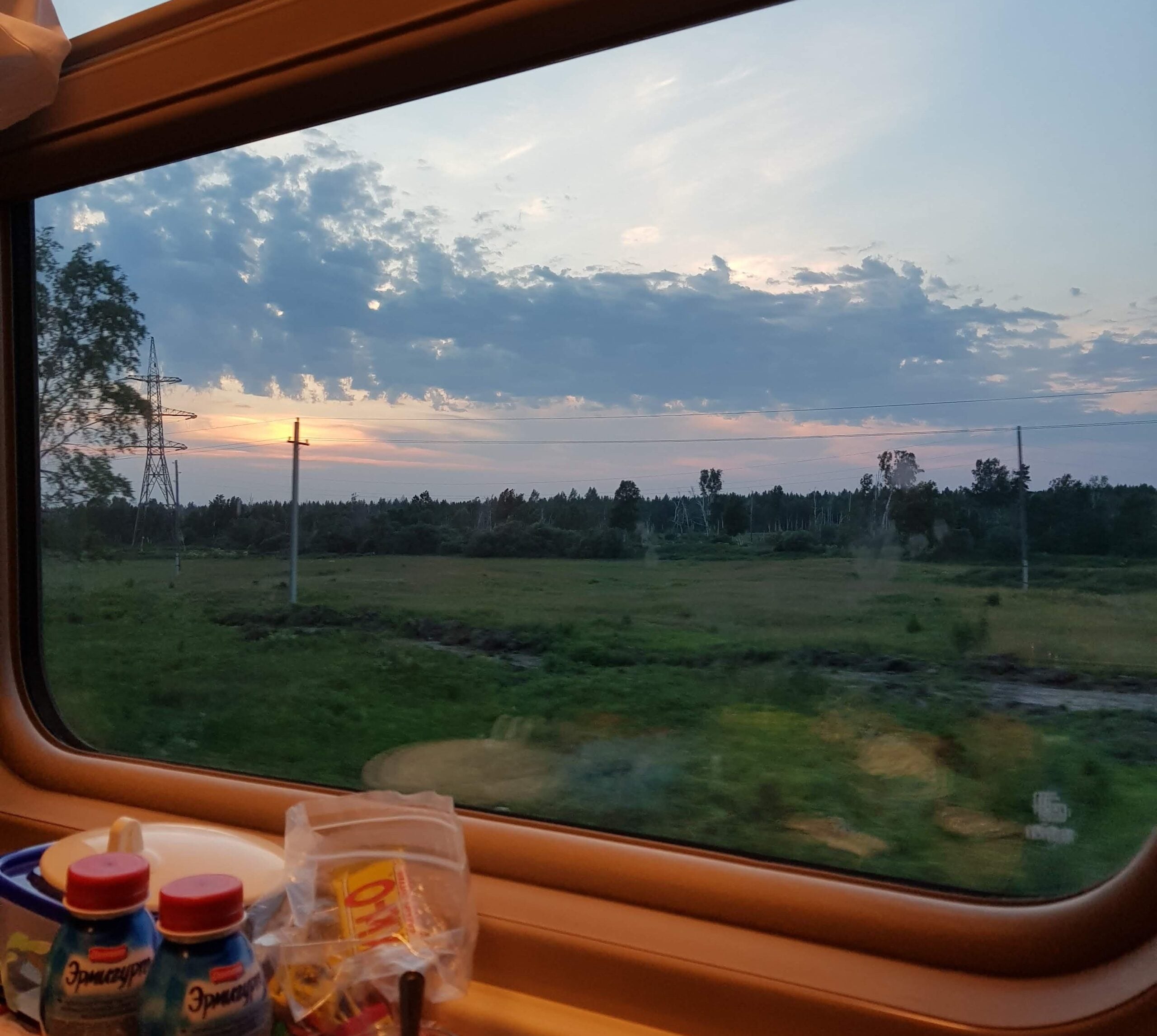View from a train window at sunset, showing green fields, trees, and a partly cloudy sky. Inside the train, snacks and drinks are visible on a table near the window.