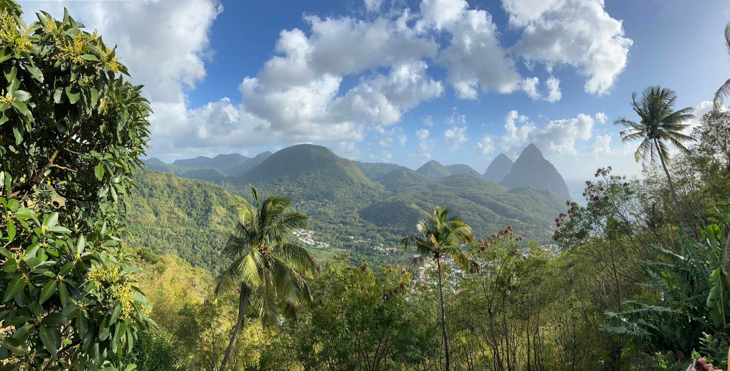 Lush green mountains with palm trees in the foreground evoke the beauty of Tropical Locations, while a small village nestles below and two distinct volcanic peaks rise under a partly cloudy blue sky.