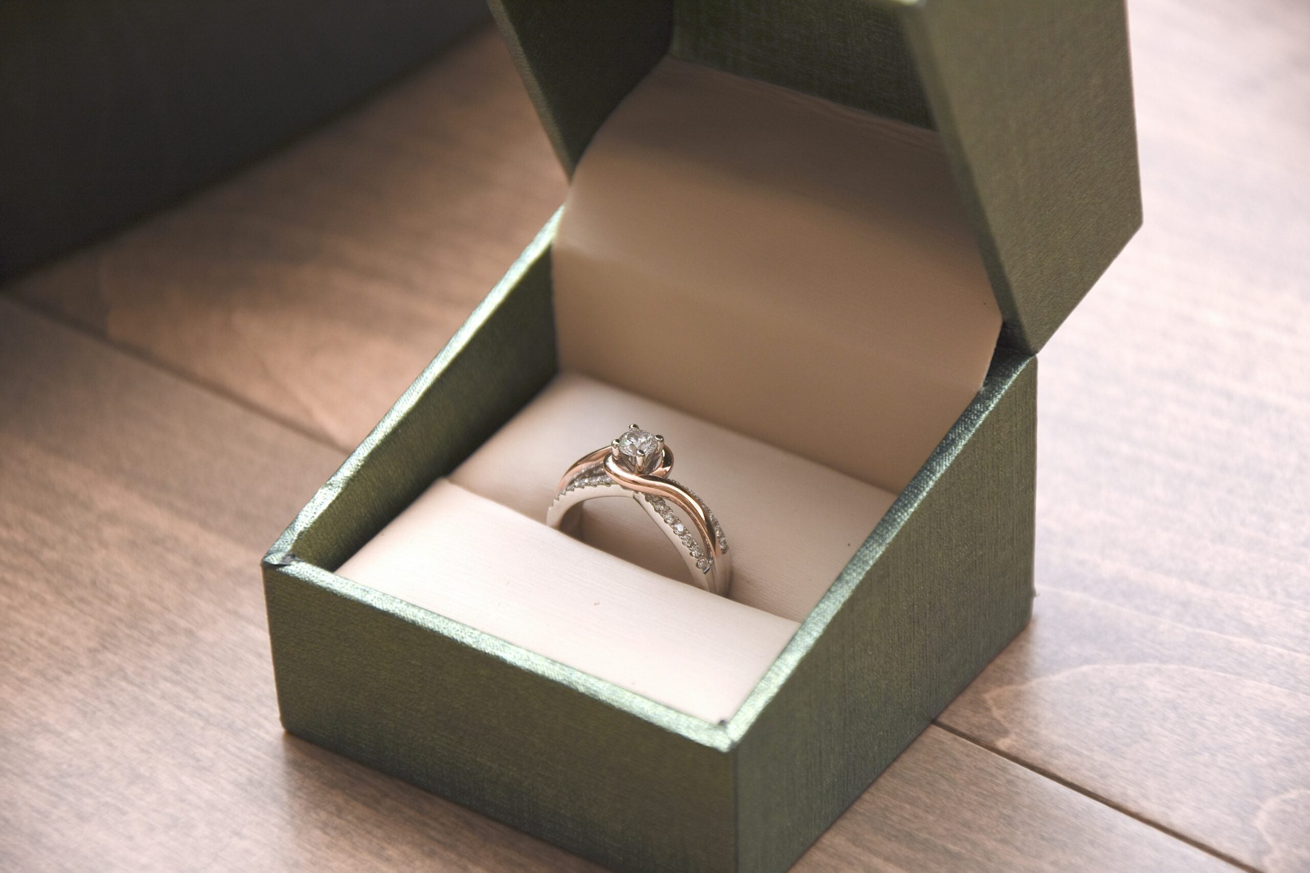 A diamond engagement ring with a twisted band is displayed in an open green box on a wooden surface.