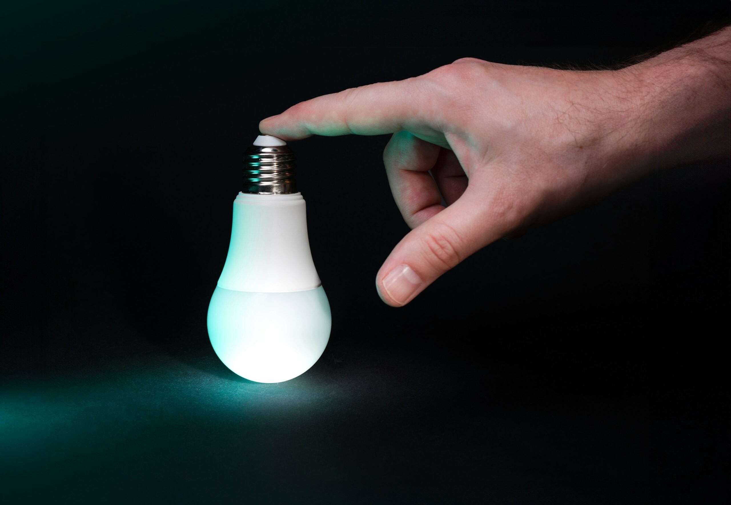 A hand gently touches the top of a glowing LED light bulb against a dark background.