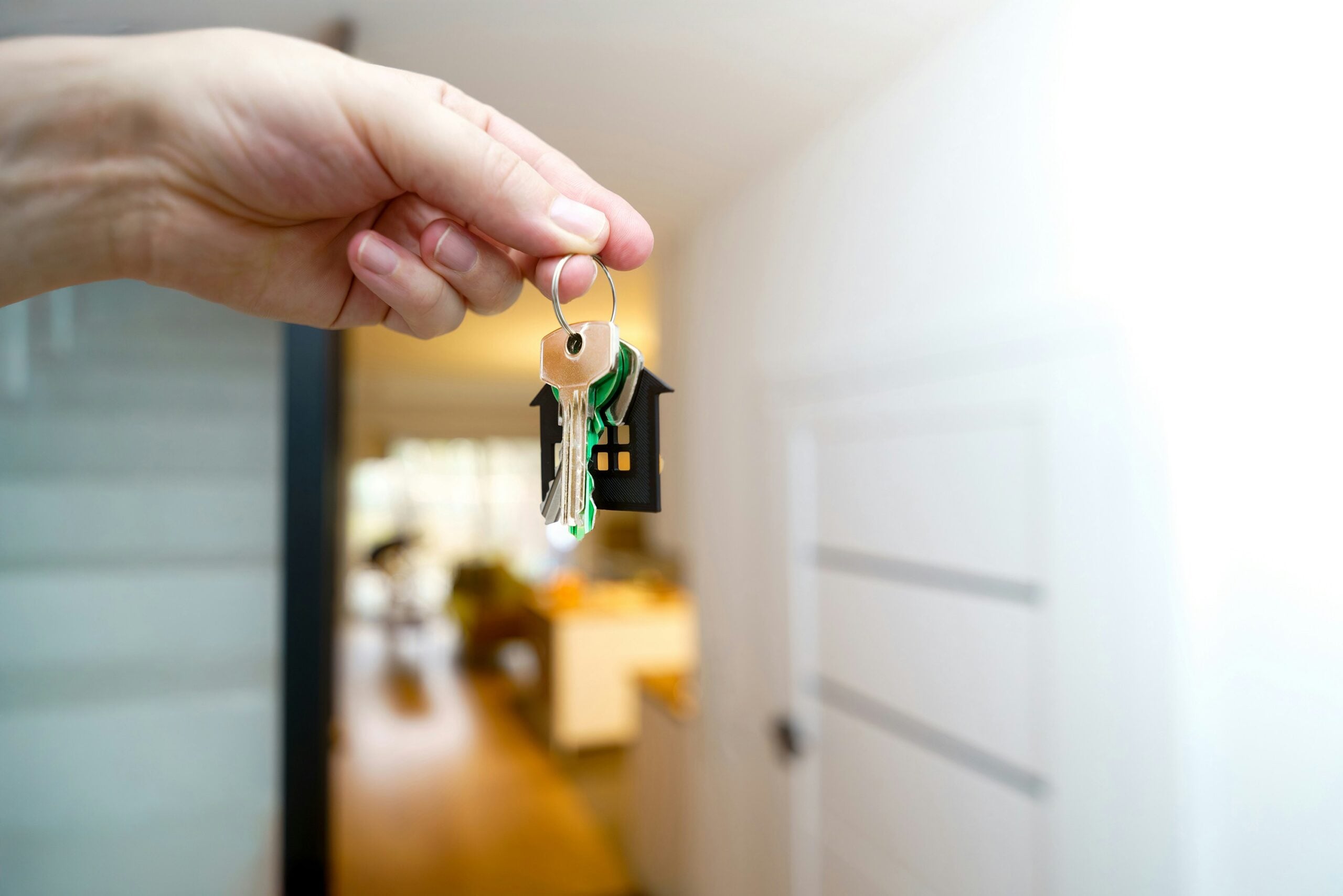 A hand holds a key ring with several keys and a small black house-shaped keychain, with a blurred home interior in the background.