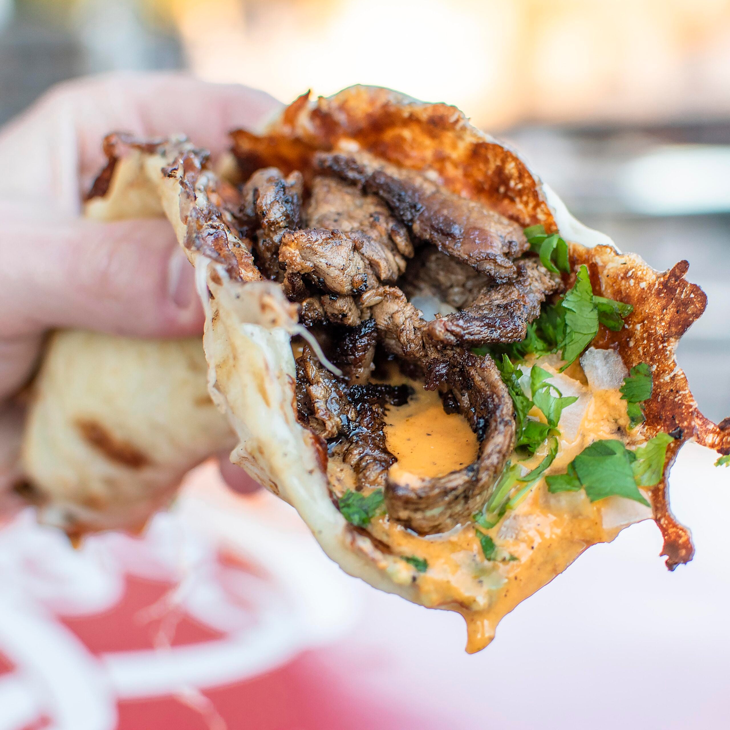 A hand holds a taco filled with grilled beef, melted cheese, creamy orange sauce, and fresh cilantro in a soft tortilla.