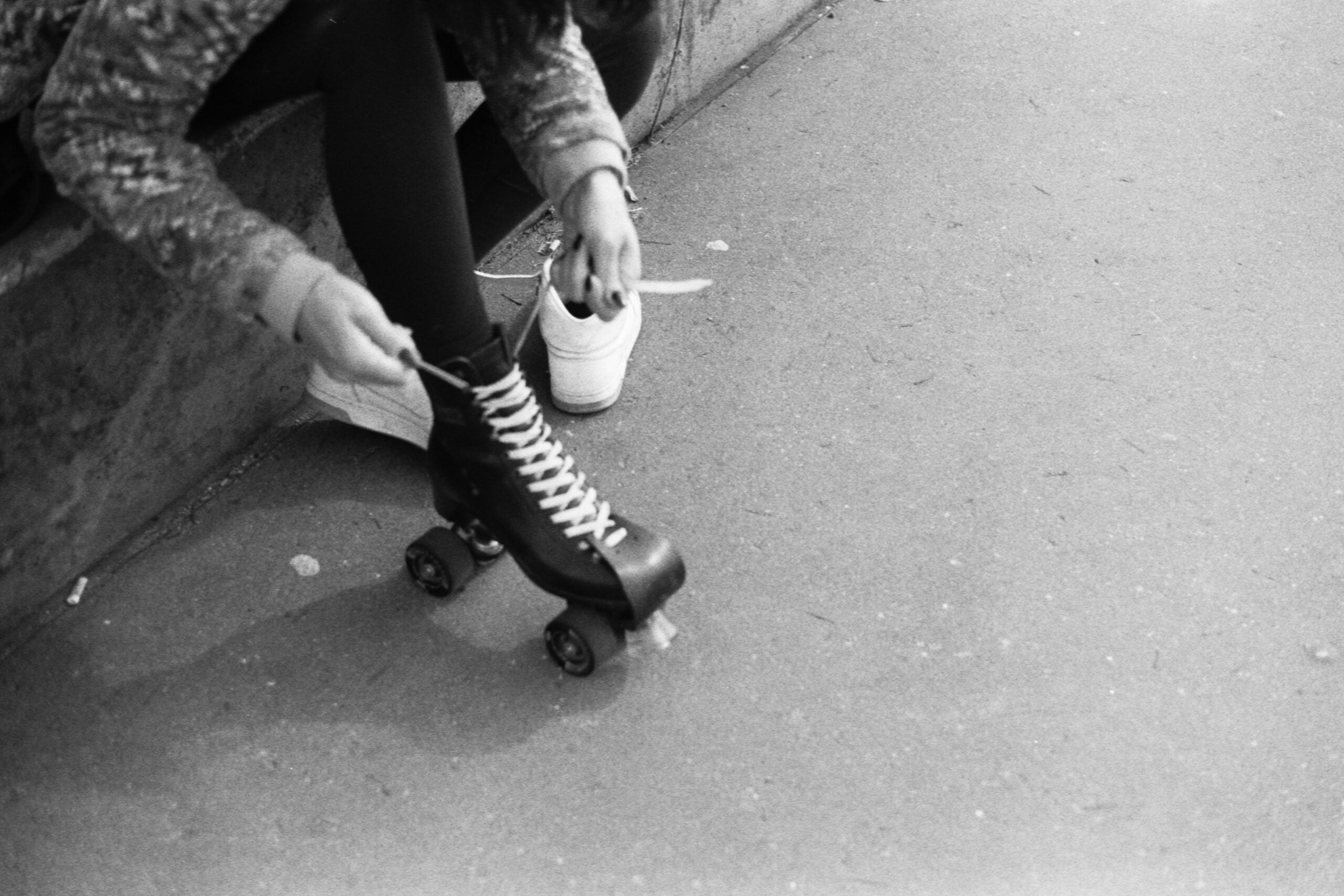 Person sitting on a curb, wearing black tights, lacing up a black roller skate on one foot while the other foot is still in a white sneaker. Photo is black and white.