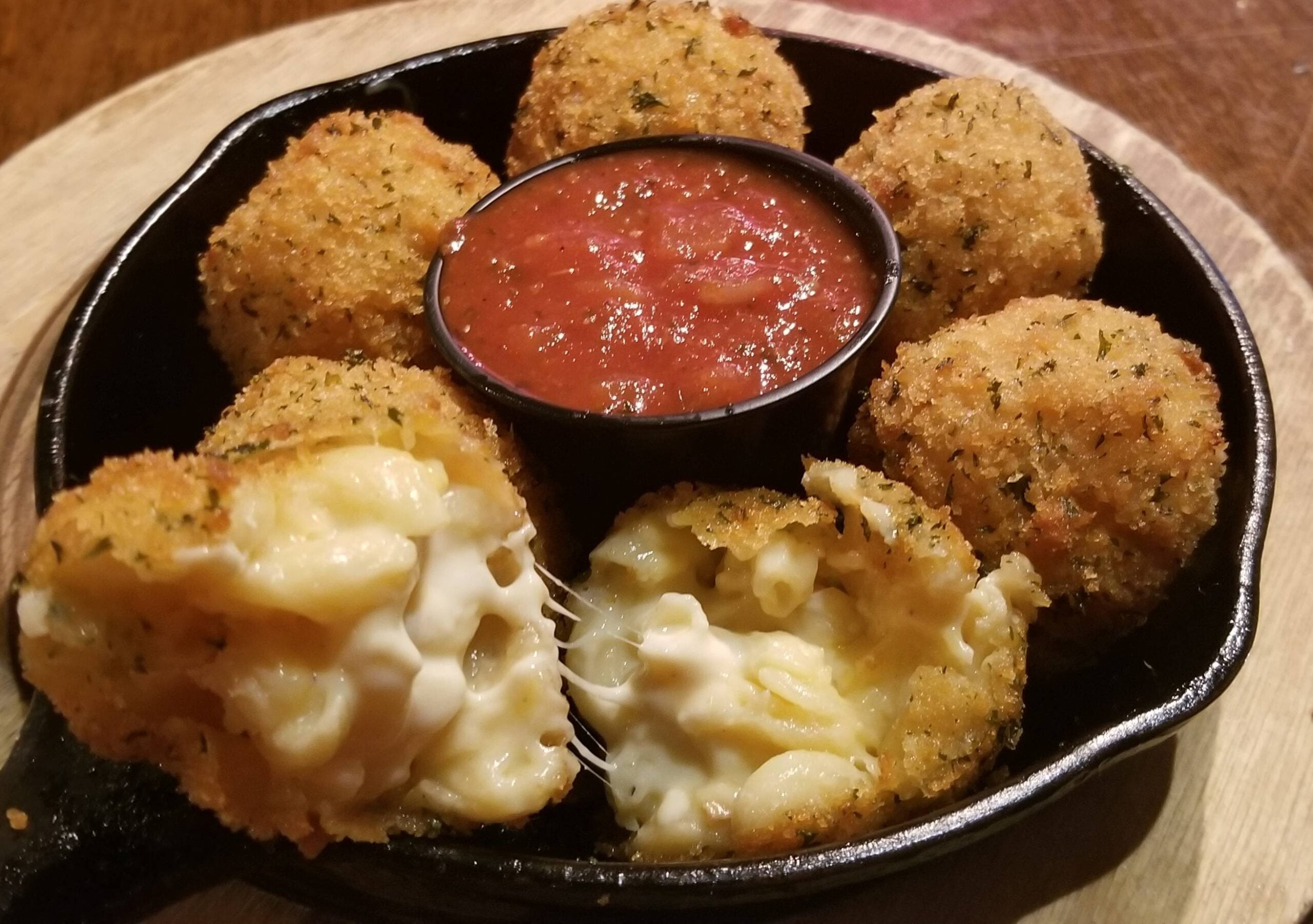 Six breaded, fried macaroni and cheese balls in a skillet with a cup of marinara sauce in the center. One ball is split open, showing the creamy mac and cheese filling.