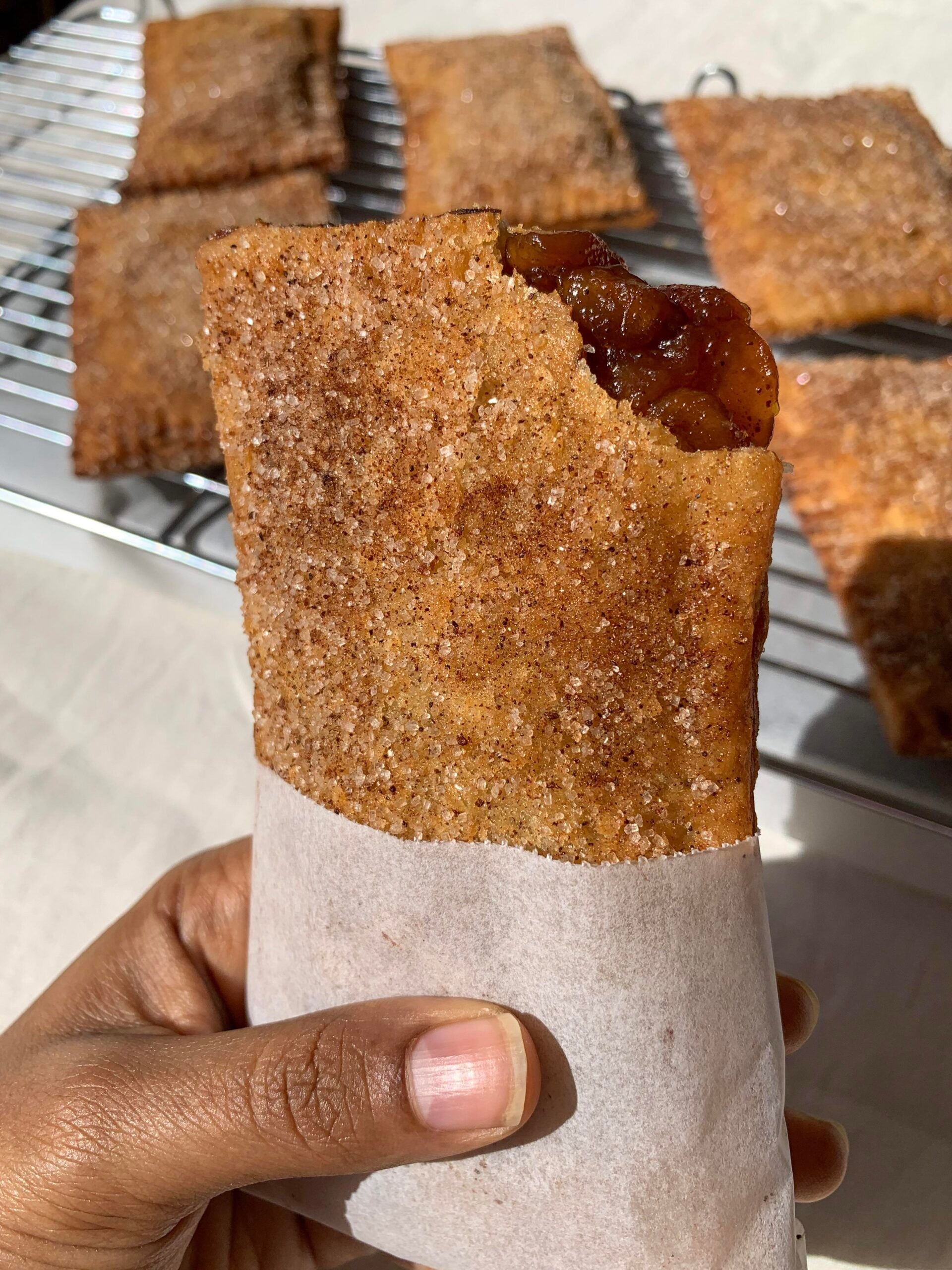 A hand holds a baked pastry filled with apple chunks, coated in cinnamon sugar, with a bite taken out. Cooling pastries are visible on a rack in the background.