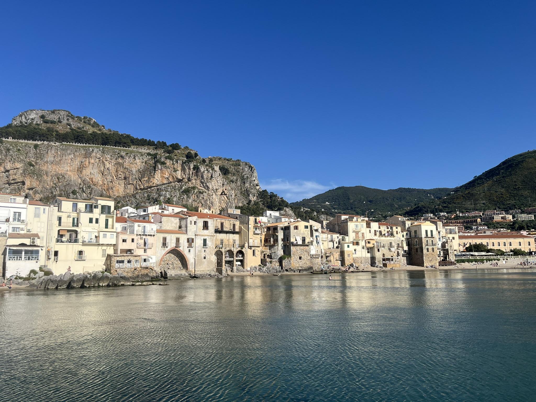 Colorful Mediterranean buildings line a calm waterfront with clear blue skies, mountains, and rocky cliffs in the background. The shoreline reflects the houses in the still water, creating a picturesque scene near some of the region’s most incredible beaches.