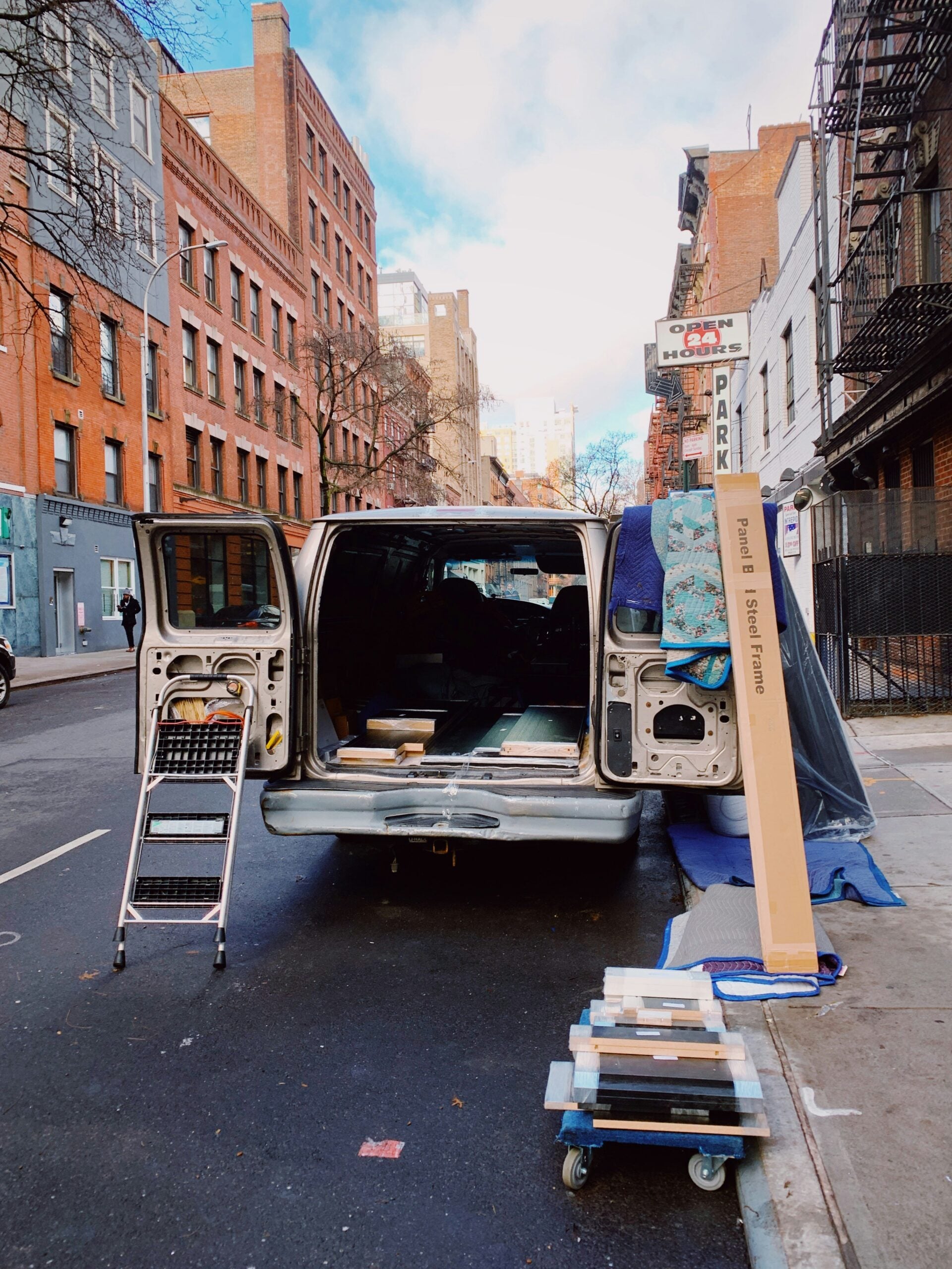 A van with open doors is parked on a city street. A step ladder, furniture, and boxed items are nearby, suggesting a move or delivery. Tall brick buildings and bare trees line the street.