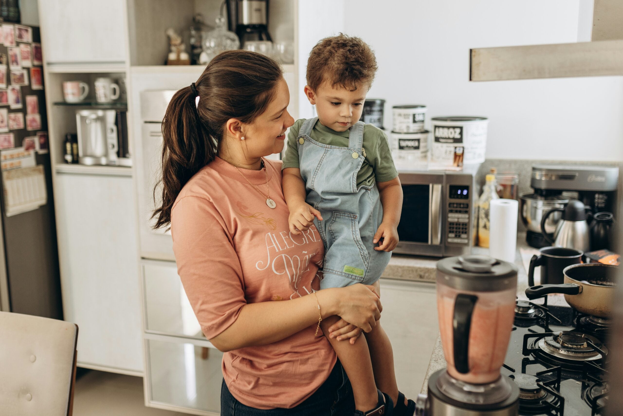 A woman holding a young child in a kitchen looks at a blender filled with a pink smoothie, with various kitchen items and appliances visible on the counters.