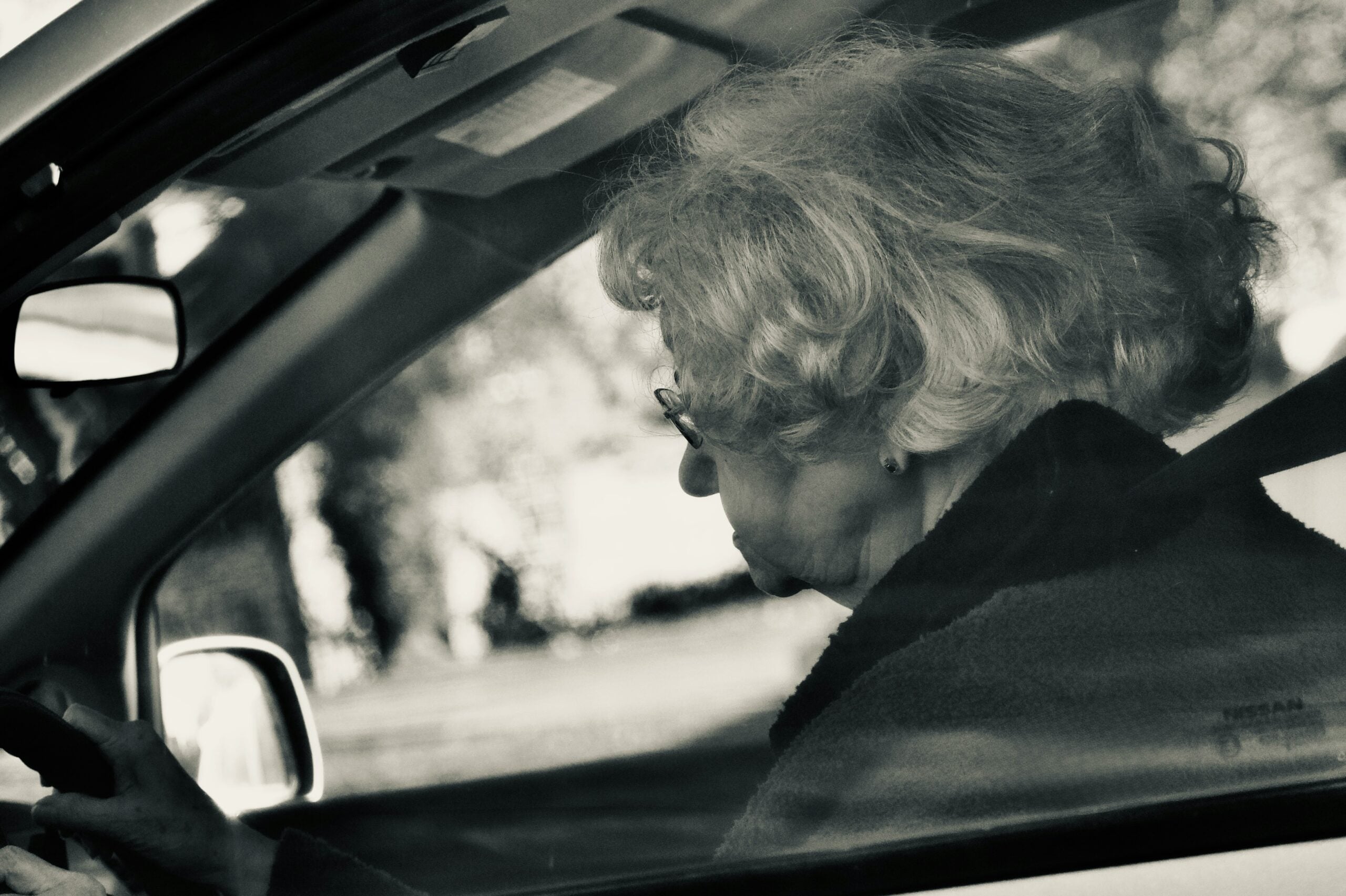 An elderly woman with glasses and curly hair is sitting in the driver’s seat of a car, looking straight ahead through the window, perhaps reflecting on her retirement savings as she heads out for the day.