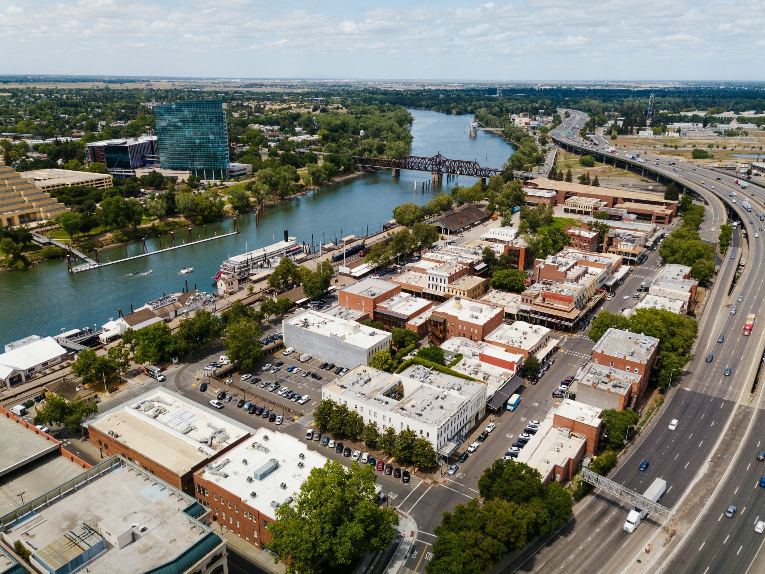 Aerial view of a city by a river, with historic brick buildings, modern glass building, highways, bridges, trees, and boats docked along the waterfront on a partly cloudy day—a vibrant scene that inspires thoughts of relaxation and planning for the future, much like building your retirement savings amid the dynamic flow of urban life.