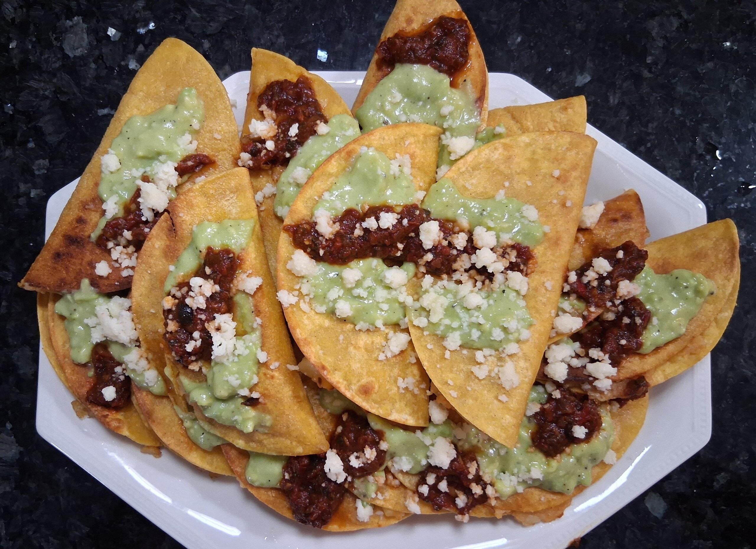 A plate of folded, fried tortillas topped with green sauce, red salsa, and crumbled white cheese, arranged on a white dish over a dark countertop.