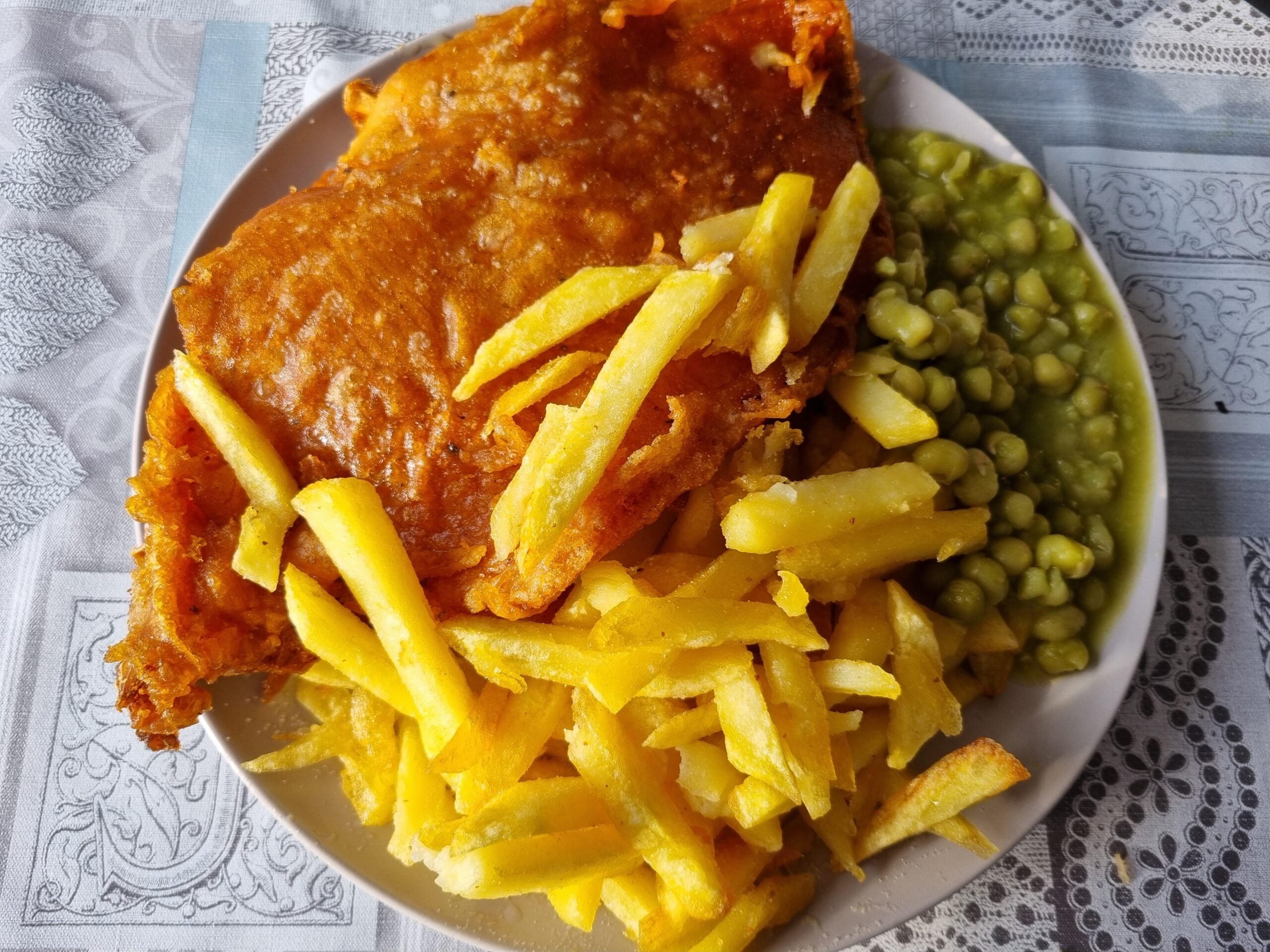 A plate with battered fried fish, golden fries, and a serving of mushy peas sits on a table with a lace-patterned tablecloth.