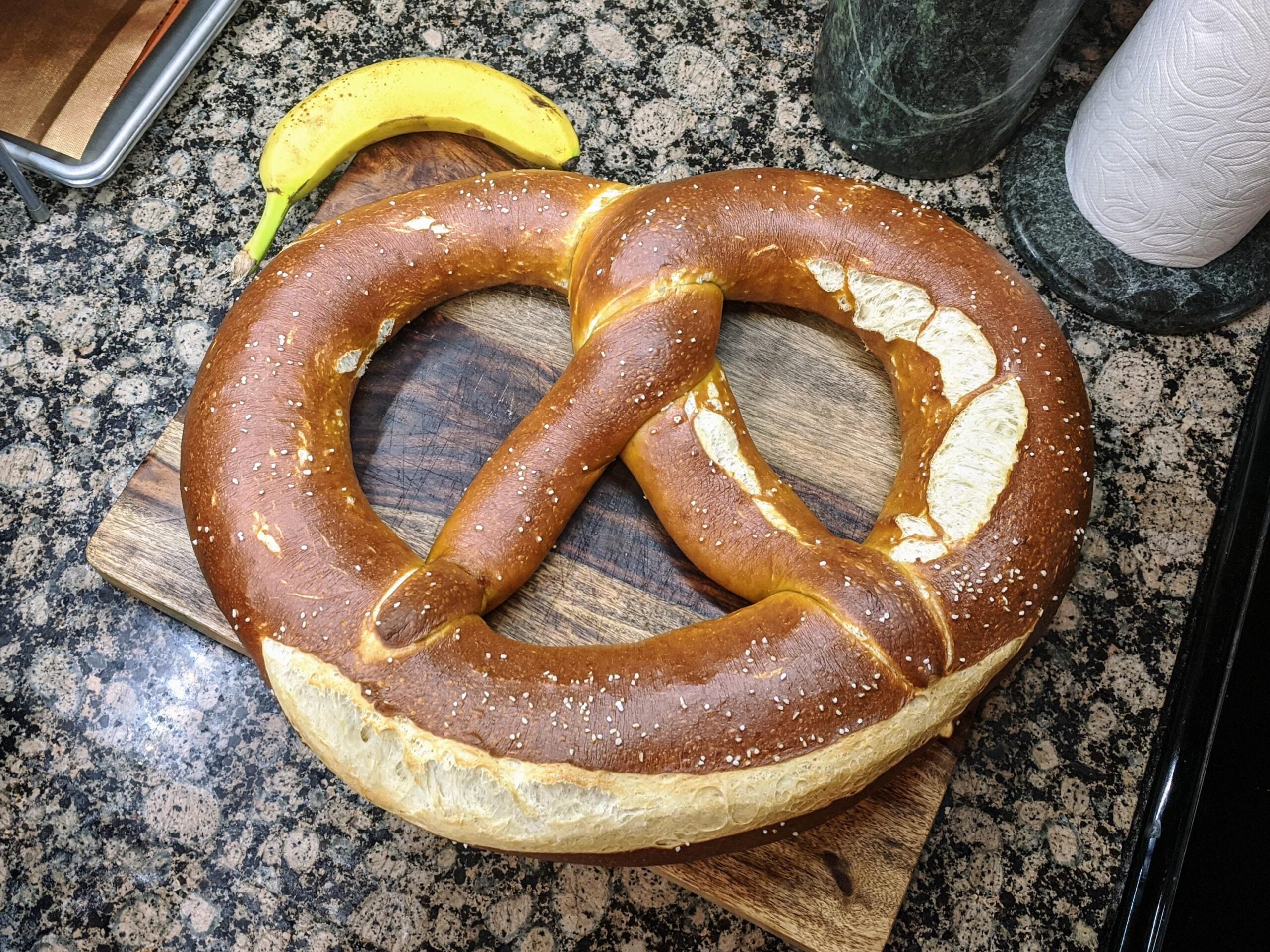 One of the giant pretzels with salt sits on a wooden cutting board next to a banana, on a granite countertop.
