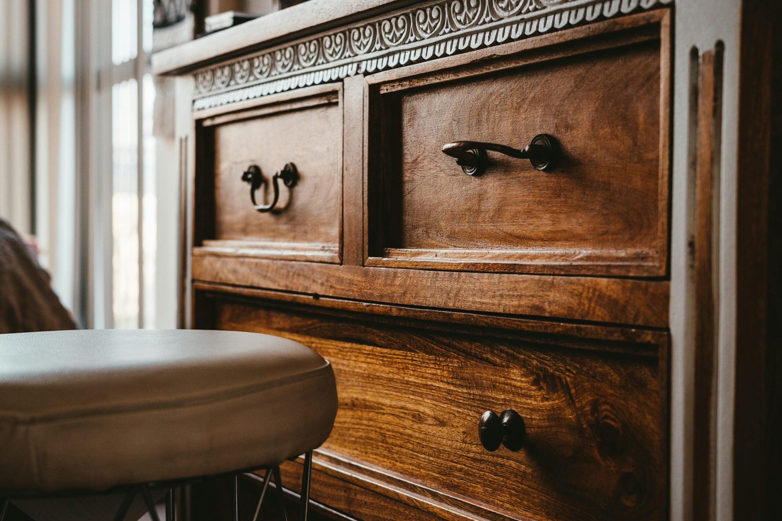 Close-up of a wooden dresser with ornate details and mismatched drawer handles, next to a round, cushioned stool. Warm sunlight streams in from a nearby window.