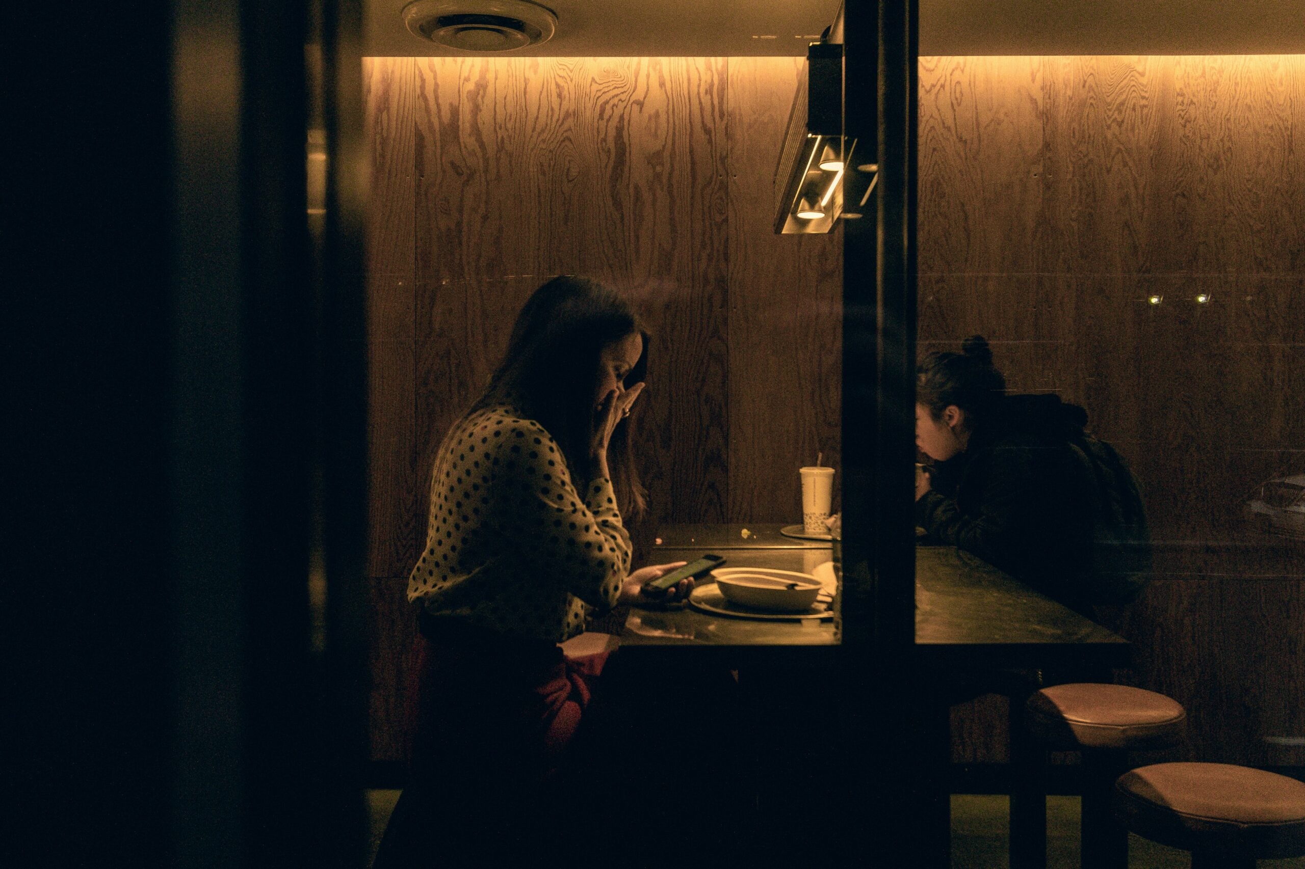 Two people sit at a dimly lit table in a restaurant; one woman covers her face with her hand, while the other looks down, both appearing deep in thought or conversation.