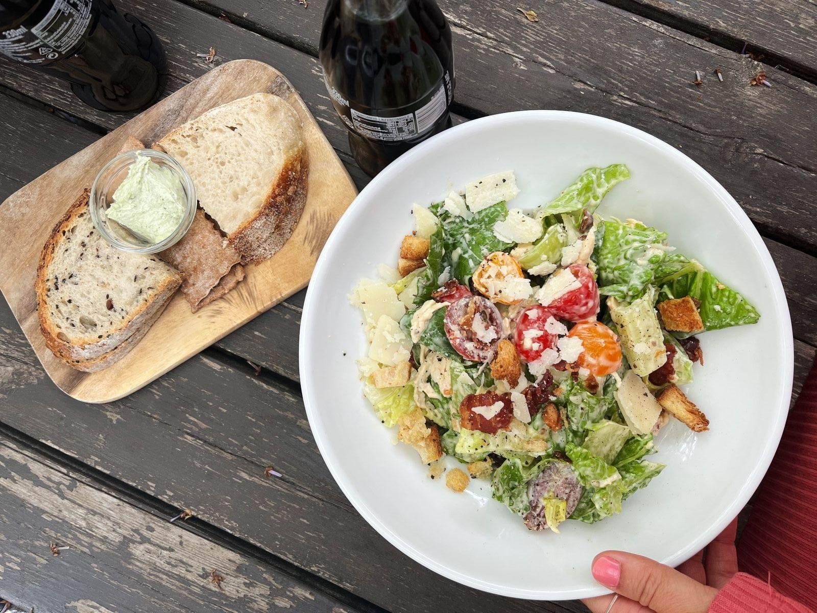 A hand holds a bowl of salad with croutons, tomatoes, and cheese. Next to it, a wooden board holds slices of bread and a small jar of spread. Two dark glass bottles are on a rustic wooden table.