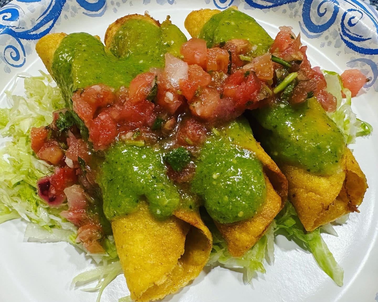 Three crispy taquitos topped with green sauce and pico de gallo, served on a bed of shredded lettuce on a decorative plate.