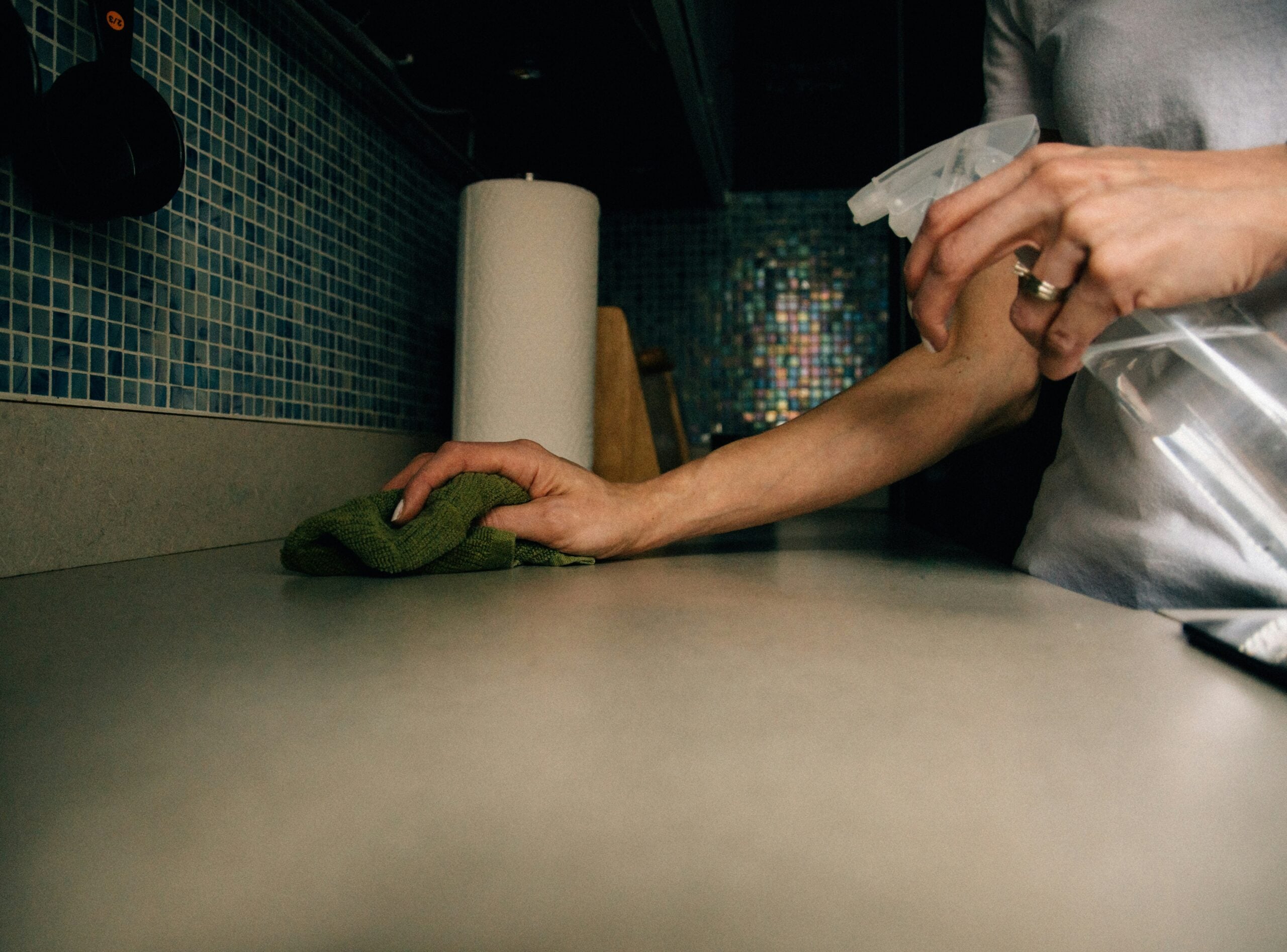 A person cleaning a kitchen counter with a green cloth in one hand and a spray bottle in the other. Blue mosaic tiles and a paper towel roll are visible in the background.