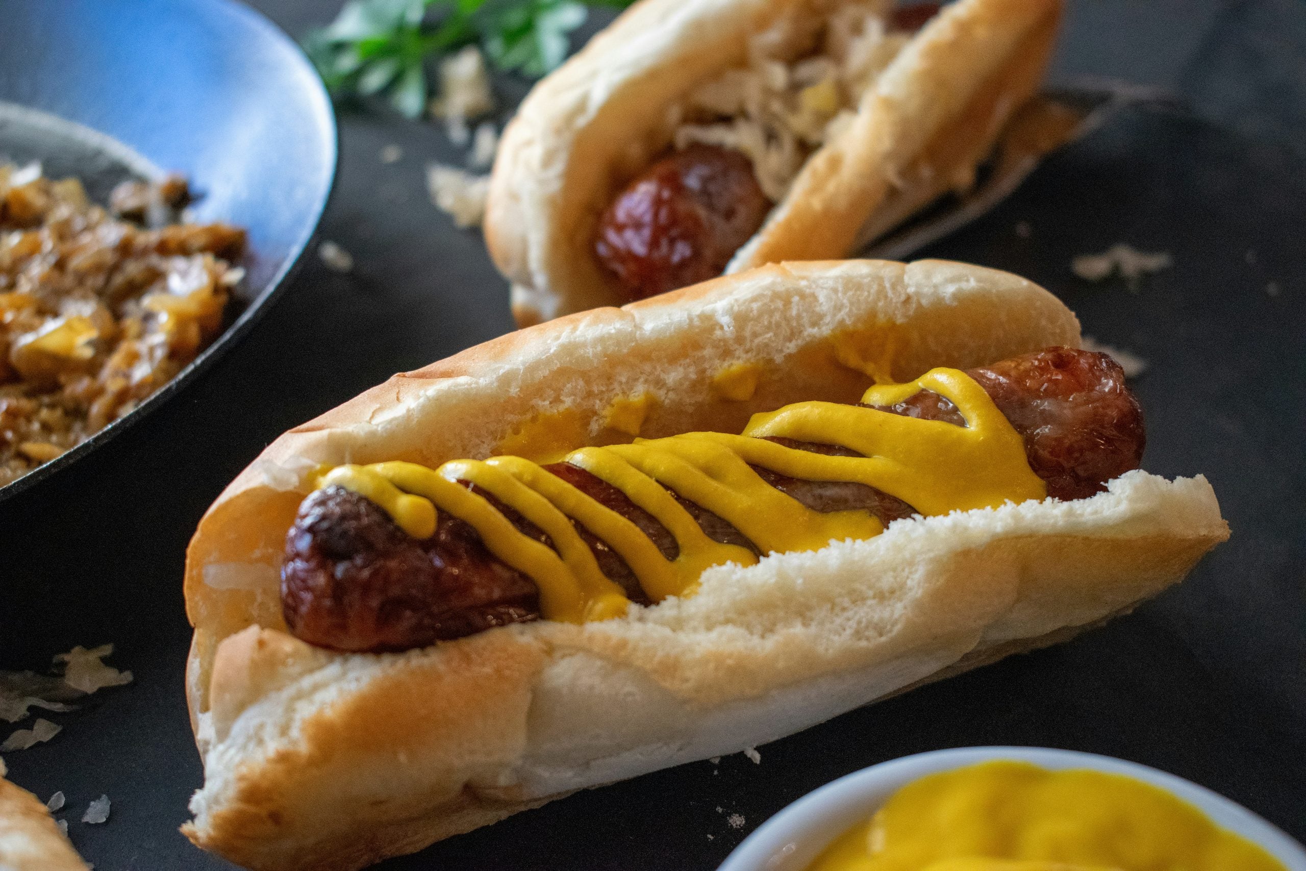 A grilled sausage in a bun topped with yellow mustard, with another sausage sandwich and a bowl of mustard in the background.