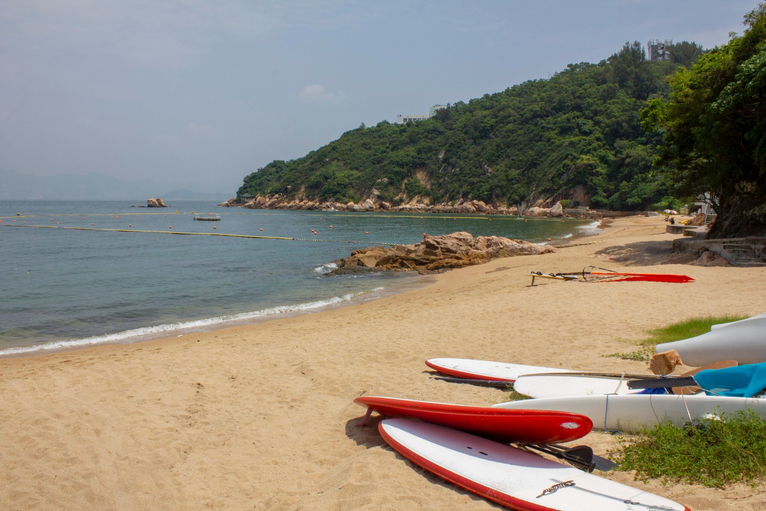 Several kayaks rest on a sandy beach beside calm water, one of the area’s incredible beaches. Green, tree-covered hills rise in the background under a lightly cloudy sky. Some floating markers are visible in the water near the shoreline.