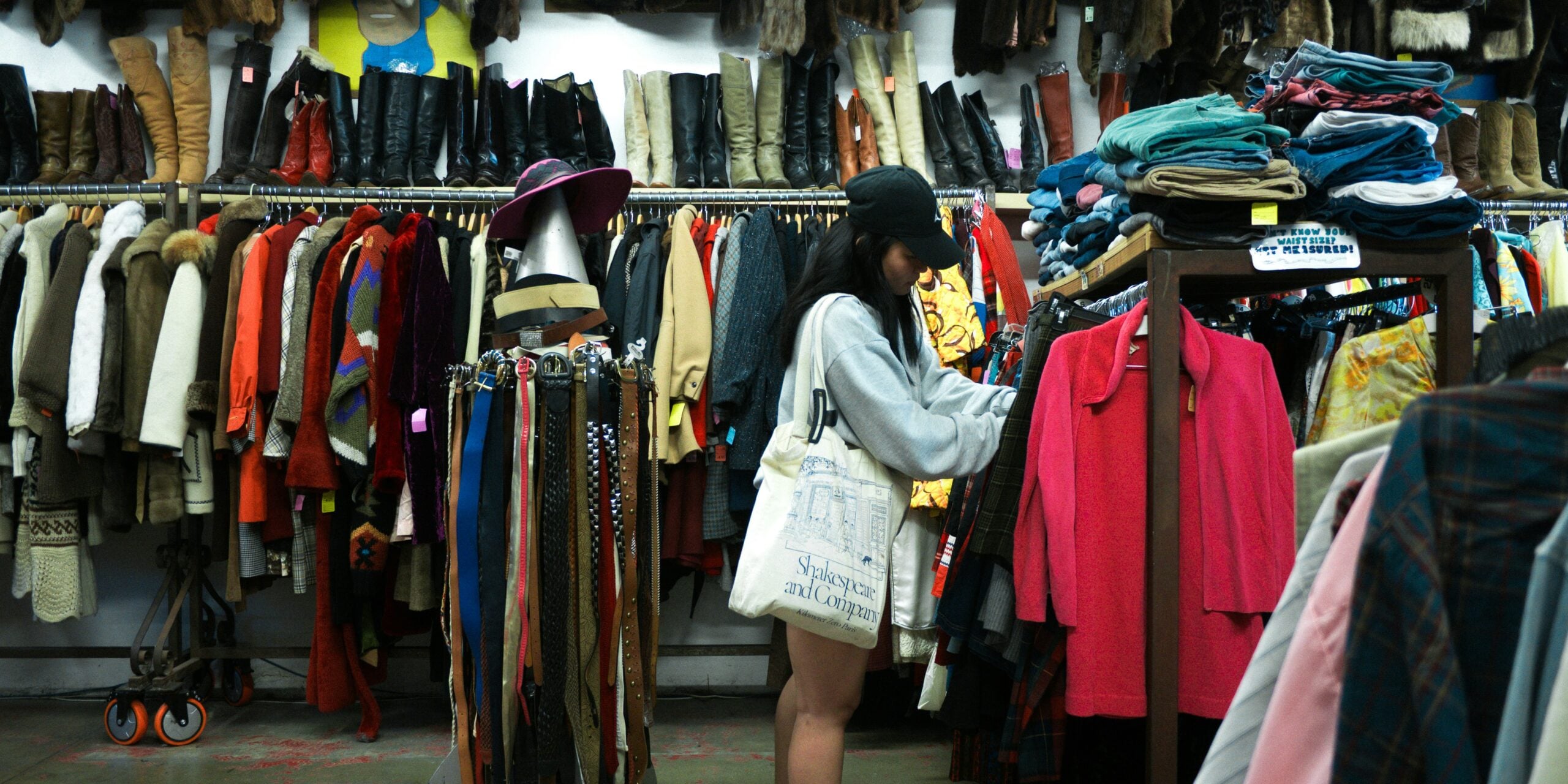 A woman browses through racks of colorful clothing in a vintage thrift store, surrounded by coats, boots, hats, and scarves organized on racks and shelves.