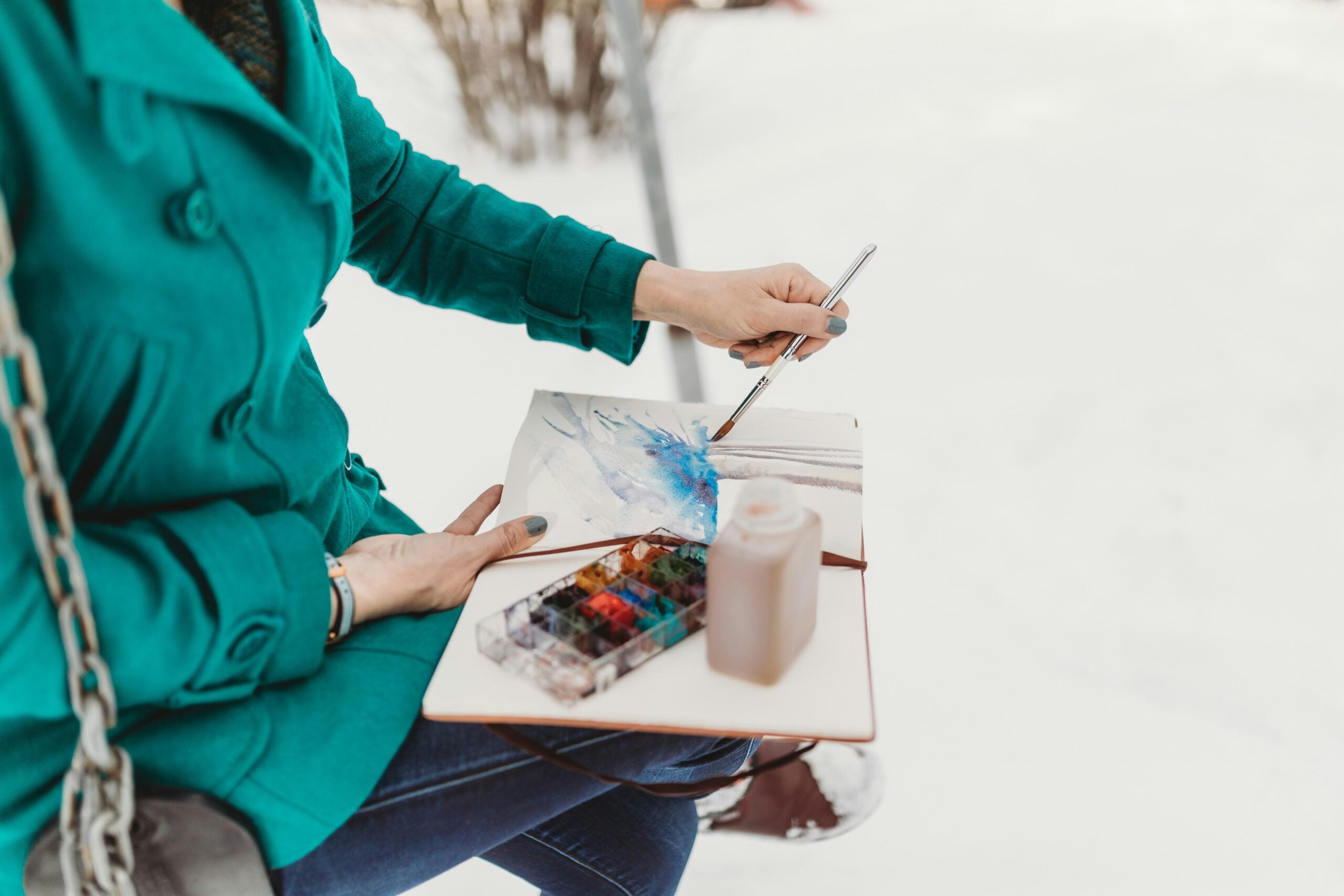 Person in a teal coat sits outside, painting a watercolor scene in a sketchbook with a brush. A paint palette and bottle are on their lap; the background is snowy.