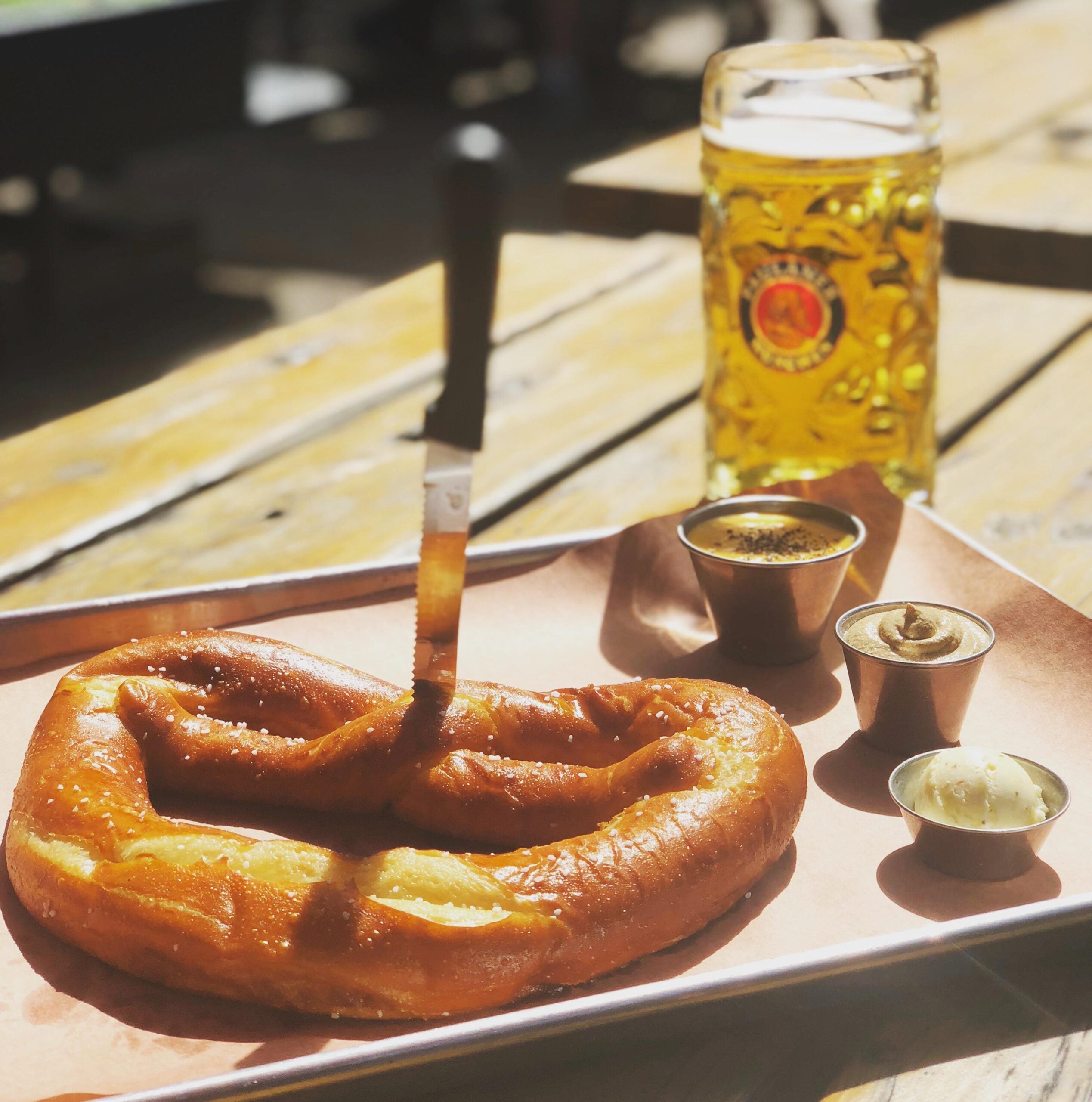 A large soft pretzel with a knife in it is served on a tray with three small cups of dips. A glass of beer sits nearby on a wooden table in bright sunlight.