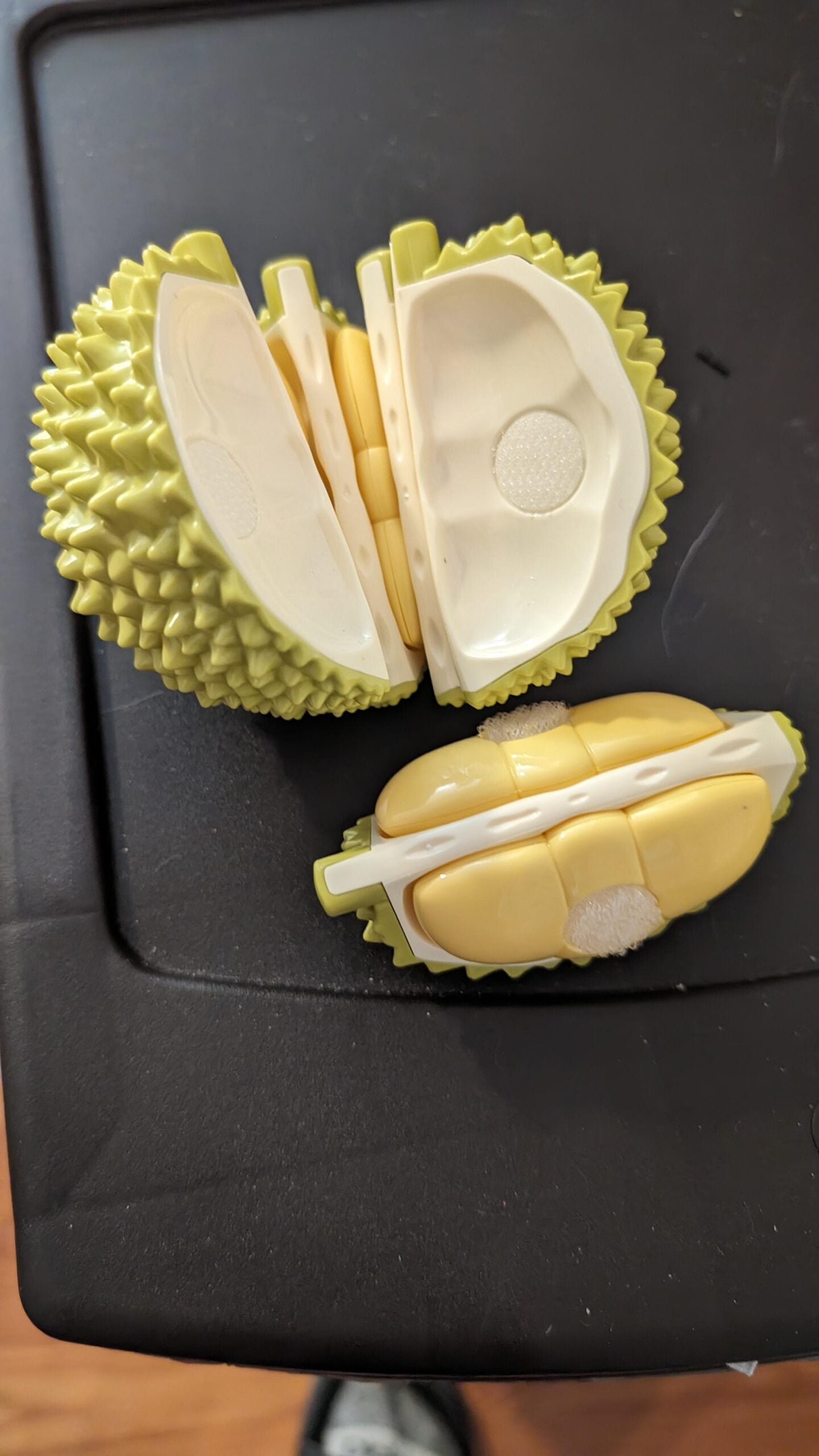 Two plastic toy durians, cut in half to reveal their interiors, rest on a black surface. The larger toy is yellow-green with white inside, the smaller one is yellow and white. Both have a spiky exterior.
