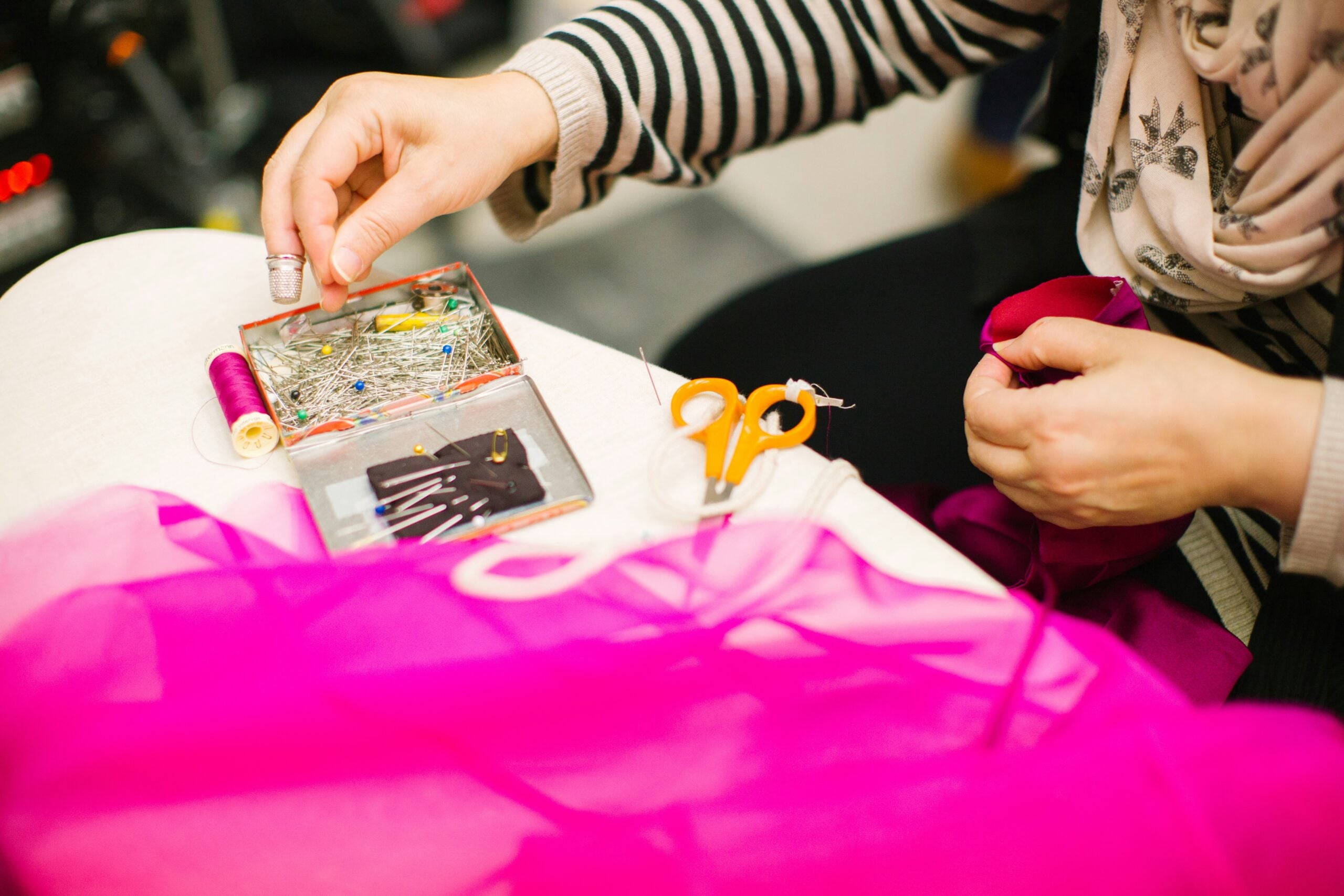 A person in a striped shirt is sewing with pink fabric, holding a needle and thread. Scissors, pins, and sewing supplies are on the table.
