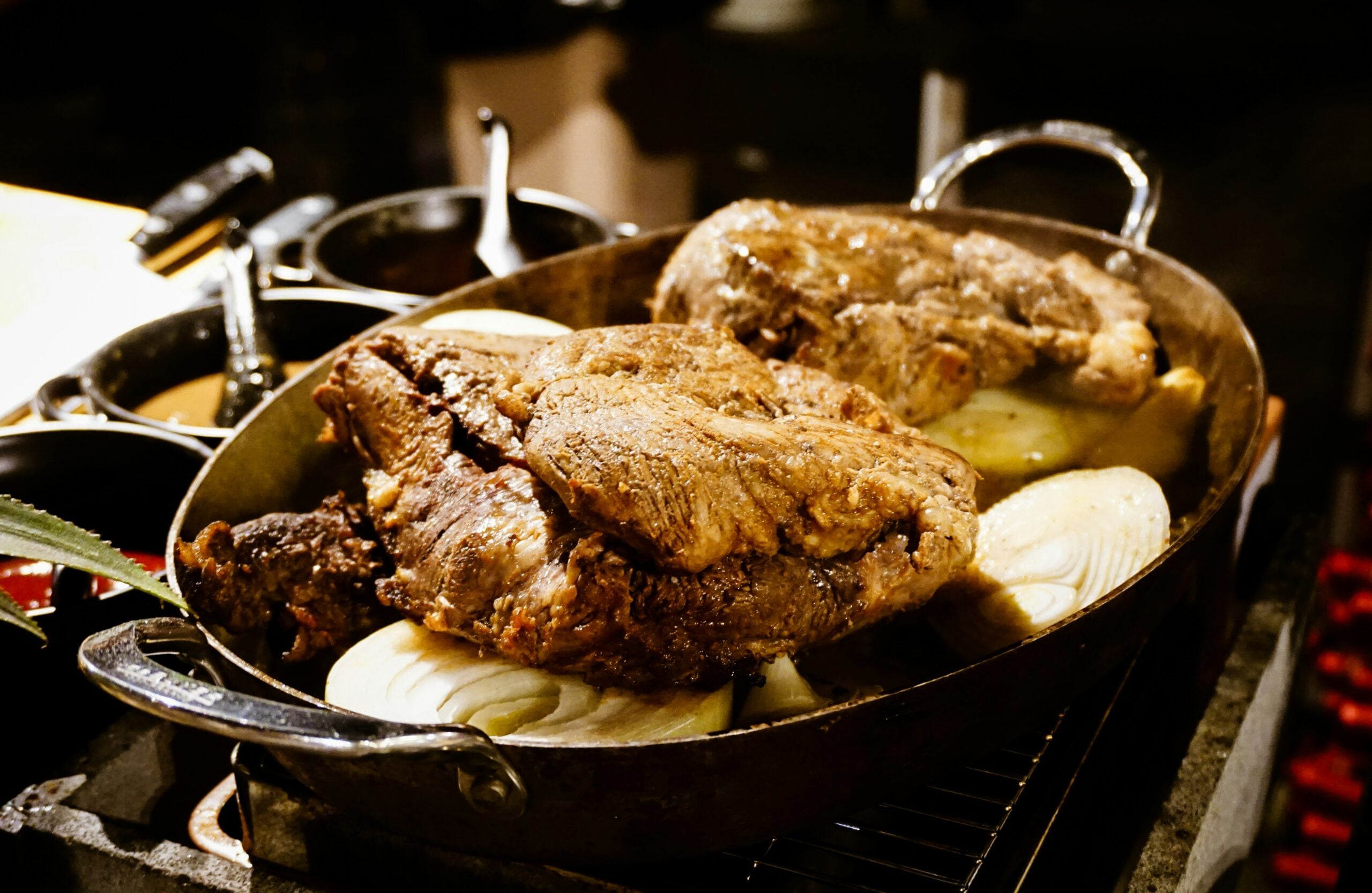 Two large pieces of roasted meat sit on top of sliced onions in a metal roasting pan, surrounded by cooking pots in a kitchen setting.