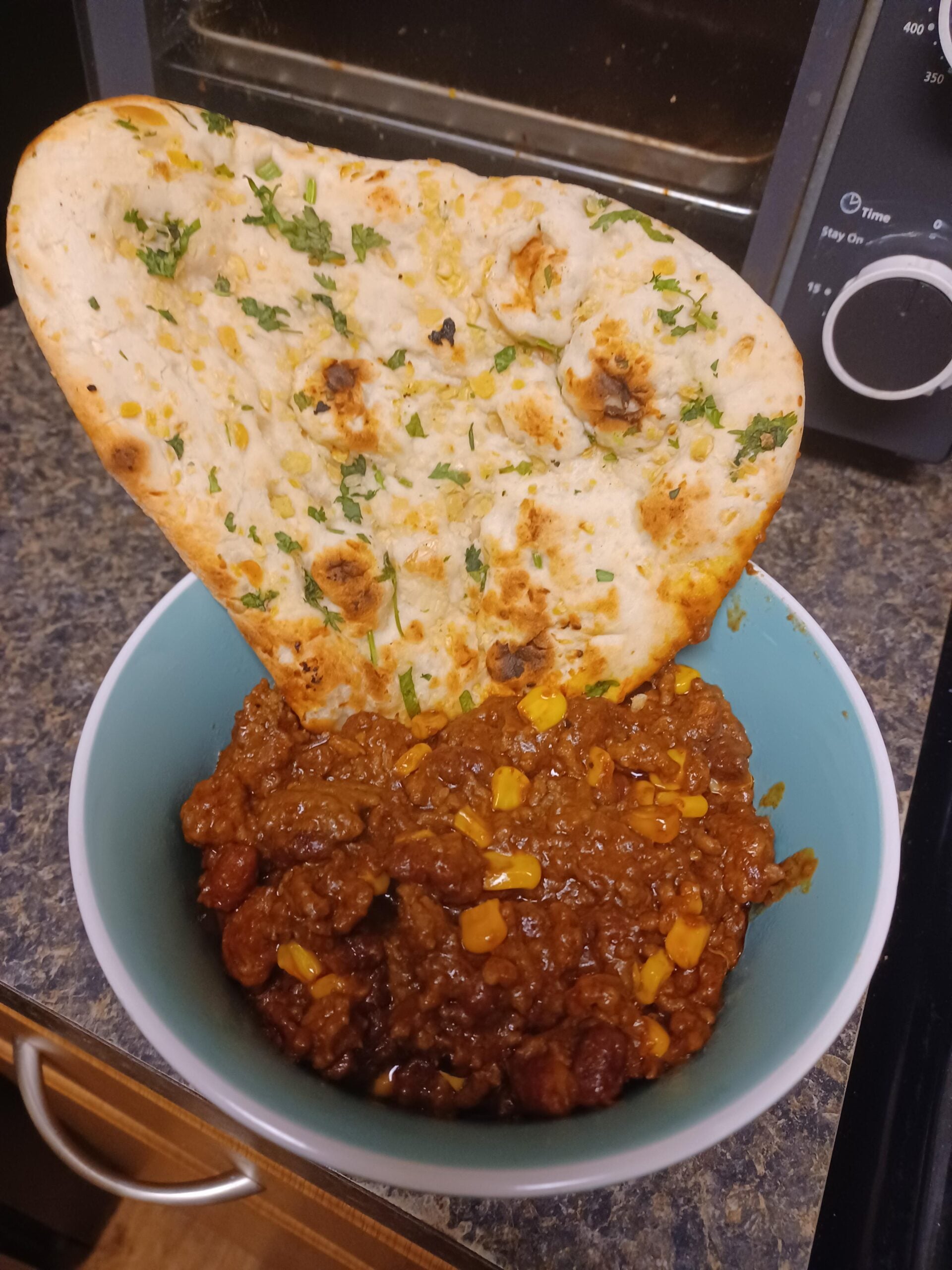 A bowl of chili with corn and beans is topped with a large piece of naan bread garnished with herbs, set on a kitchen counter in front of a toaster oven.