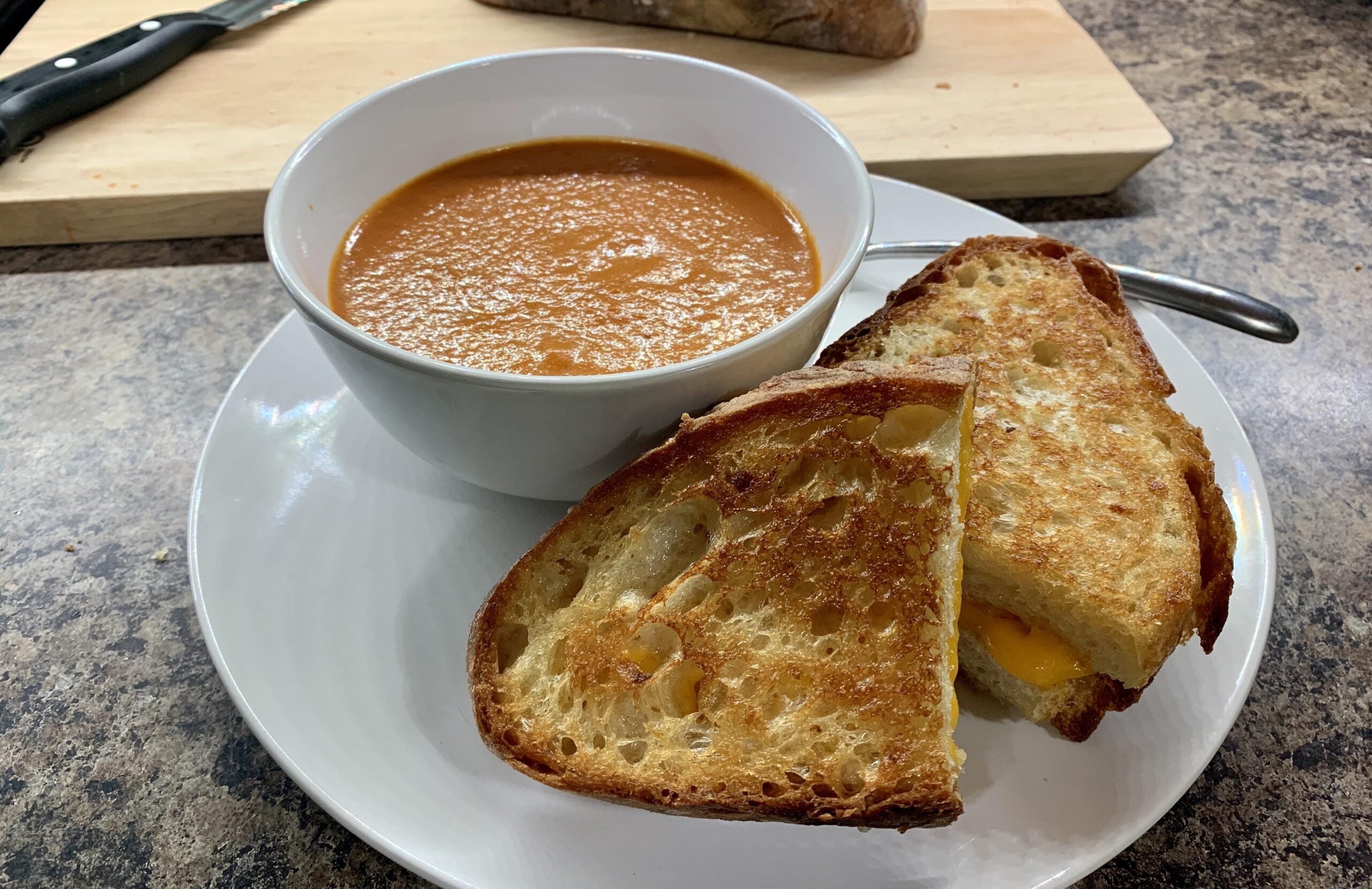 A bowl of tomato soup with a spoon sits next to one of the classic grilled cheese sandwiches on a white plate, all set on a kitchen counter.