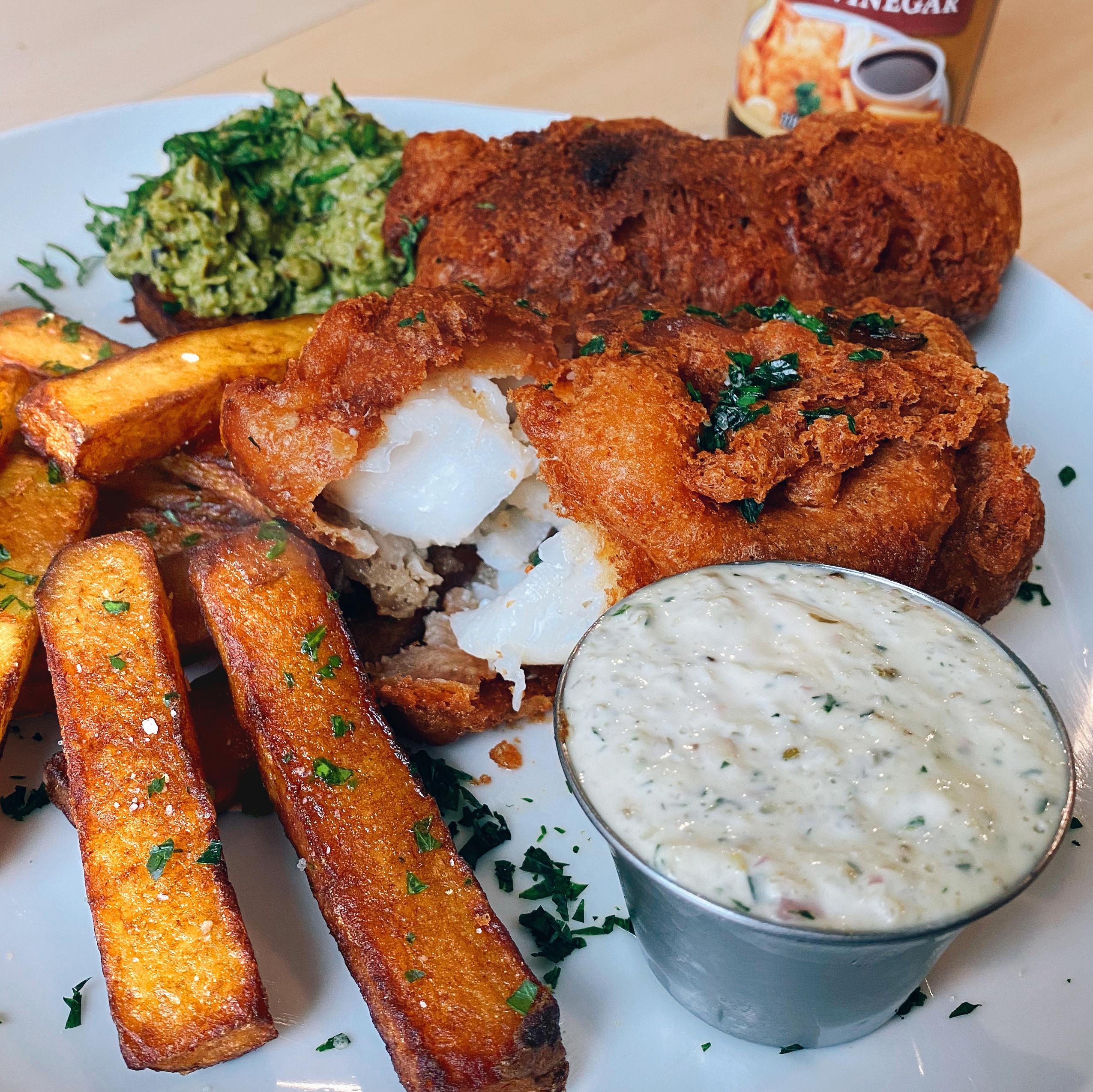A plate of golden fried fish with thick-cut fries, mushy peas, and a cup of tartar sauce. The fish is flaky inside, with crispy batter, and everything is garnished with chopped herbs.