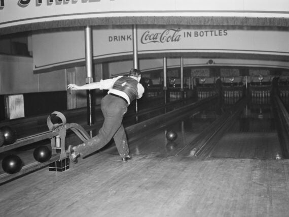 A man in a vest bowls a ball down a vintage bowling lane. Several black bowling balls sit to the side. A Coca-Cola advertisement is visible above the lanes.