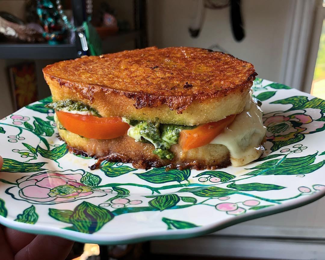 A golden-brown grilled cheese sandwich with melted cheese, tomato slices, and green pesto sits on a decorative floral plate.