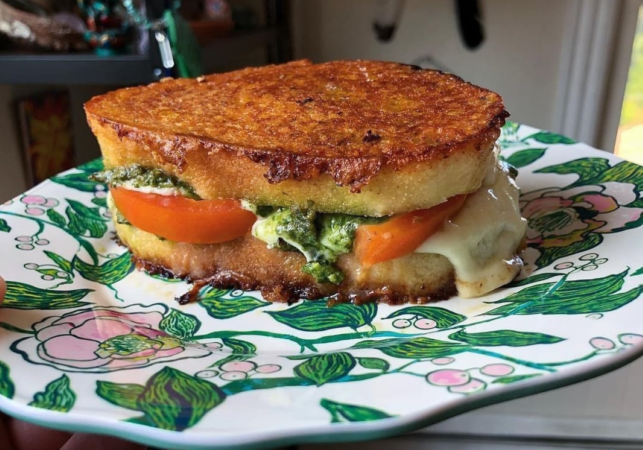 A close-up of a grilled cheese sandwich with pesto, tomato, and melted cheese on thick golden bread, served on a floral-patterned plate.