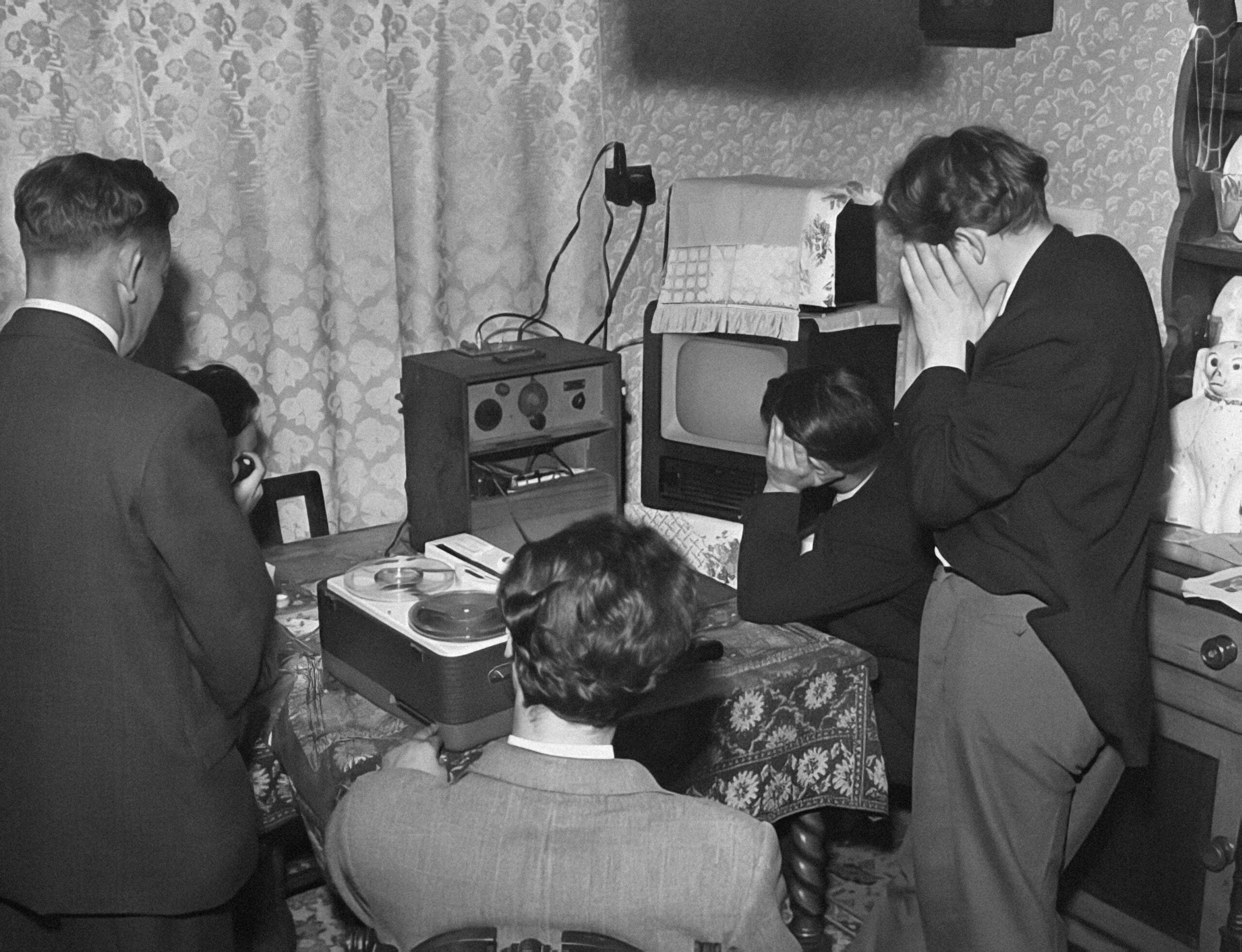 Five people in a room, some covering their faces, sit or stand around a table with a reel-to-reel tape recorder, radio, and TV. The mood appears tense or emotional. The room is decorated with patterned wallpaper and curtains.