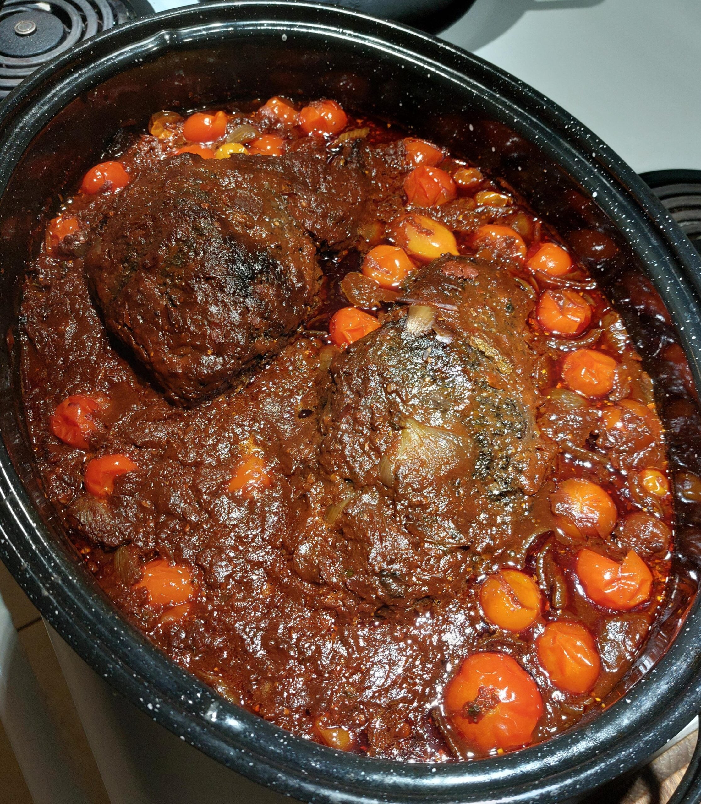 A roasting pan filled with cooked beef roasts in a rich tomato-based sauce with cherry tomatoes, set on a stovetop.
